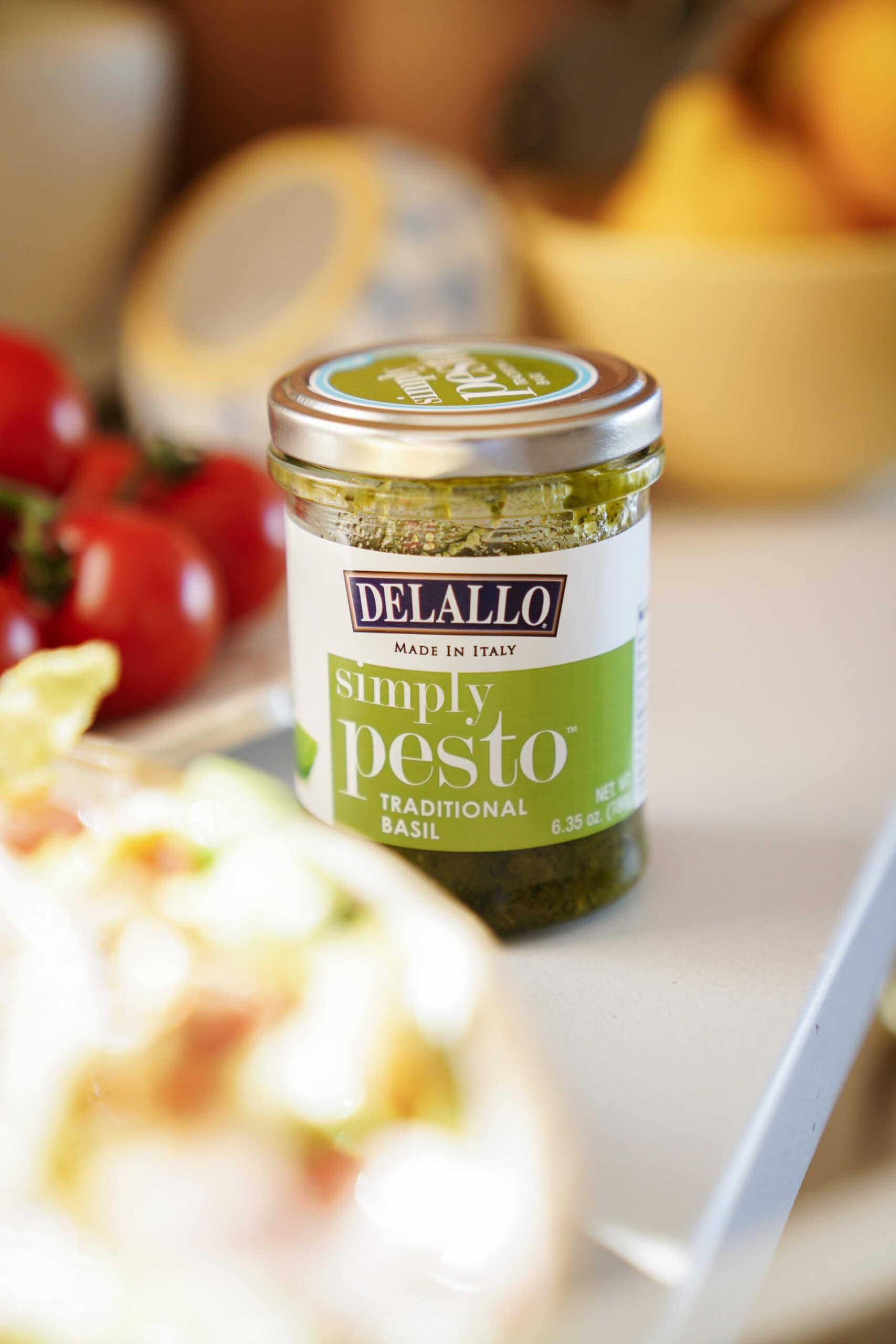 A jar of Delallo Simply Pesto Traditional Basil sits on a kitchen counter, surrounded by blurred fresh tomatoes and other food items in the background.