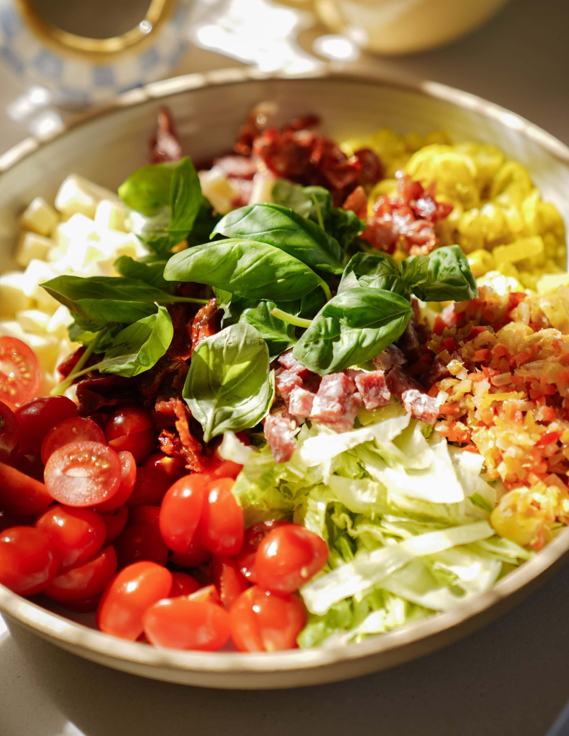 A bowl filled with chopped ingredients for an Italian salad, including cherry tomatoes, salami, lettuce, sun-dried tomatoes, cheese cubes, pepperoncini, bacon bits, and fresh basil leaves.