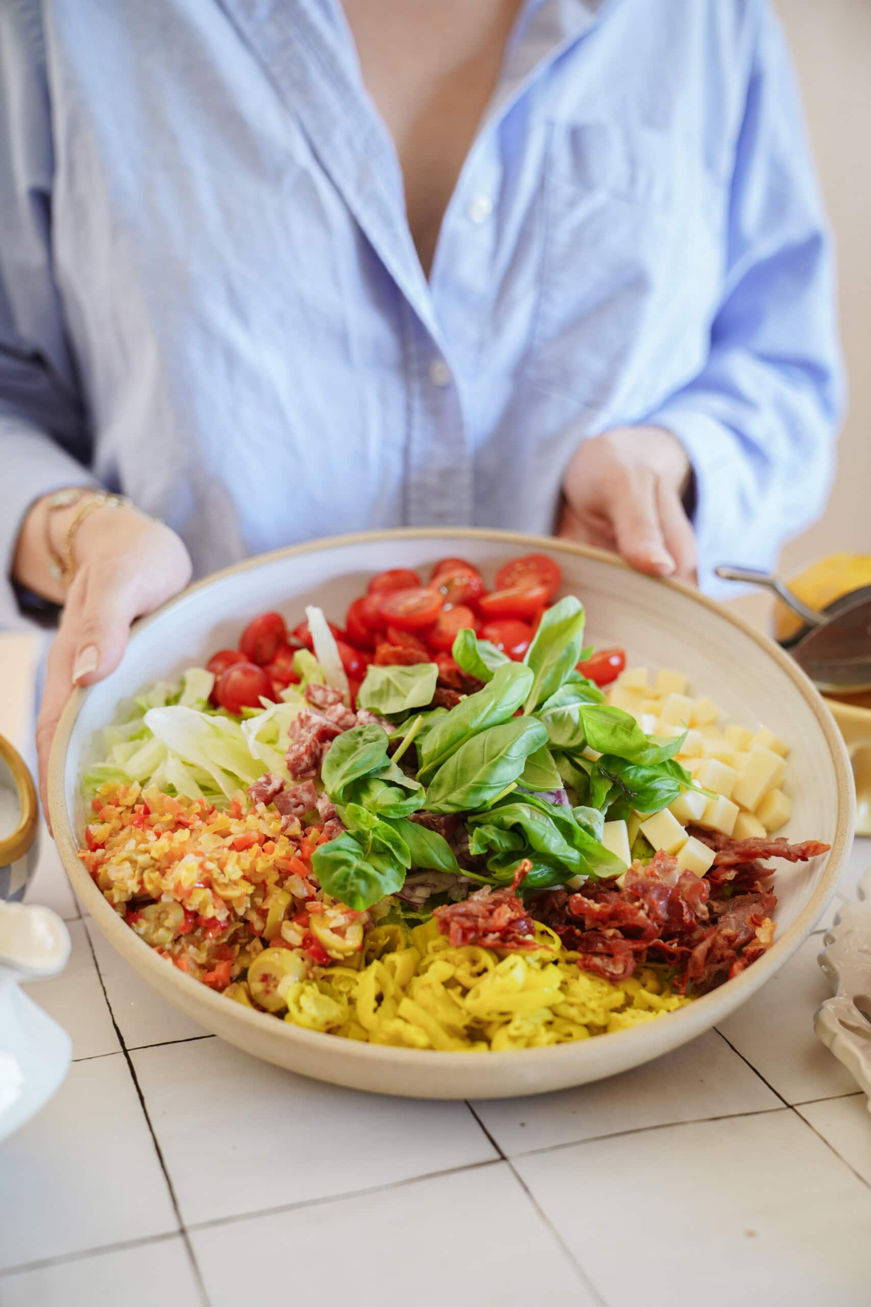 A person in a light blue shirt holds a large bowl filled with various salad ingredients, including cherry tomatoes, cheese cubes, chopped lettuce, diced meats, and fresh basil leaves.
