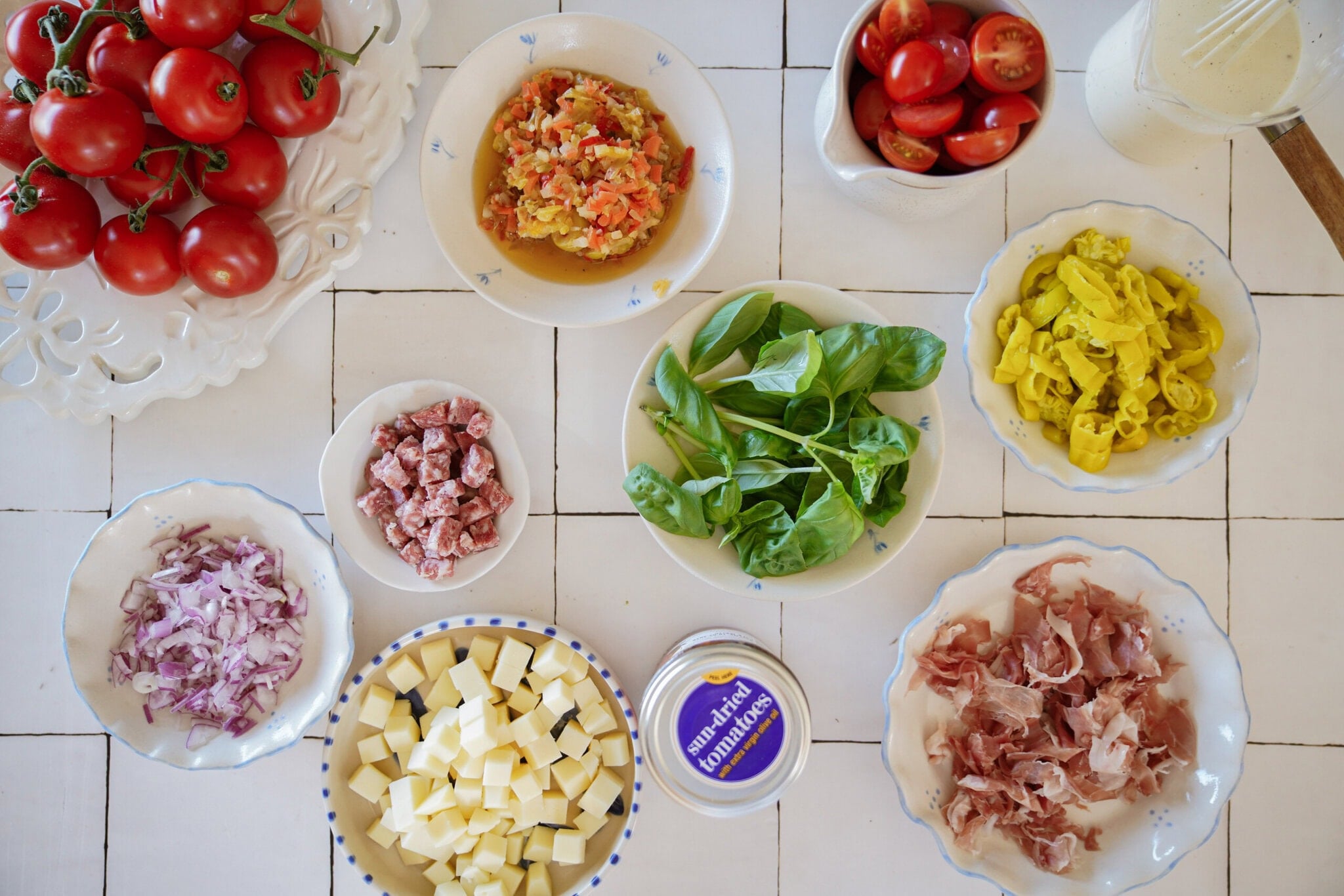 An overhead view of bowls and plates filled with fresh ingredients: tomatoes, chopped onions, cubed cheese, Italian meats, leafy greens, sliced yellow peppers, chopped mixed vegetables, and a container of mascarpone on a white tiled surface.