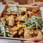 A tray of roasted chicken thighs, crispy potatoes, and green beans coated in a creamy sauce, garnished with lemon slices. The dish is being held on a parchment-lined baking sheet.