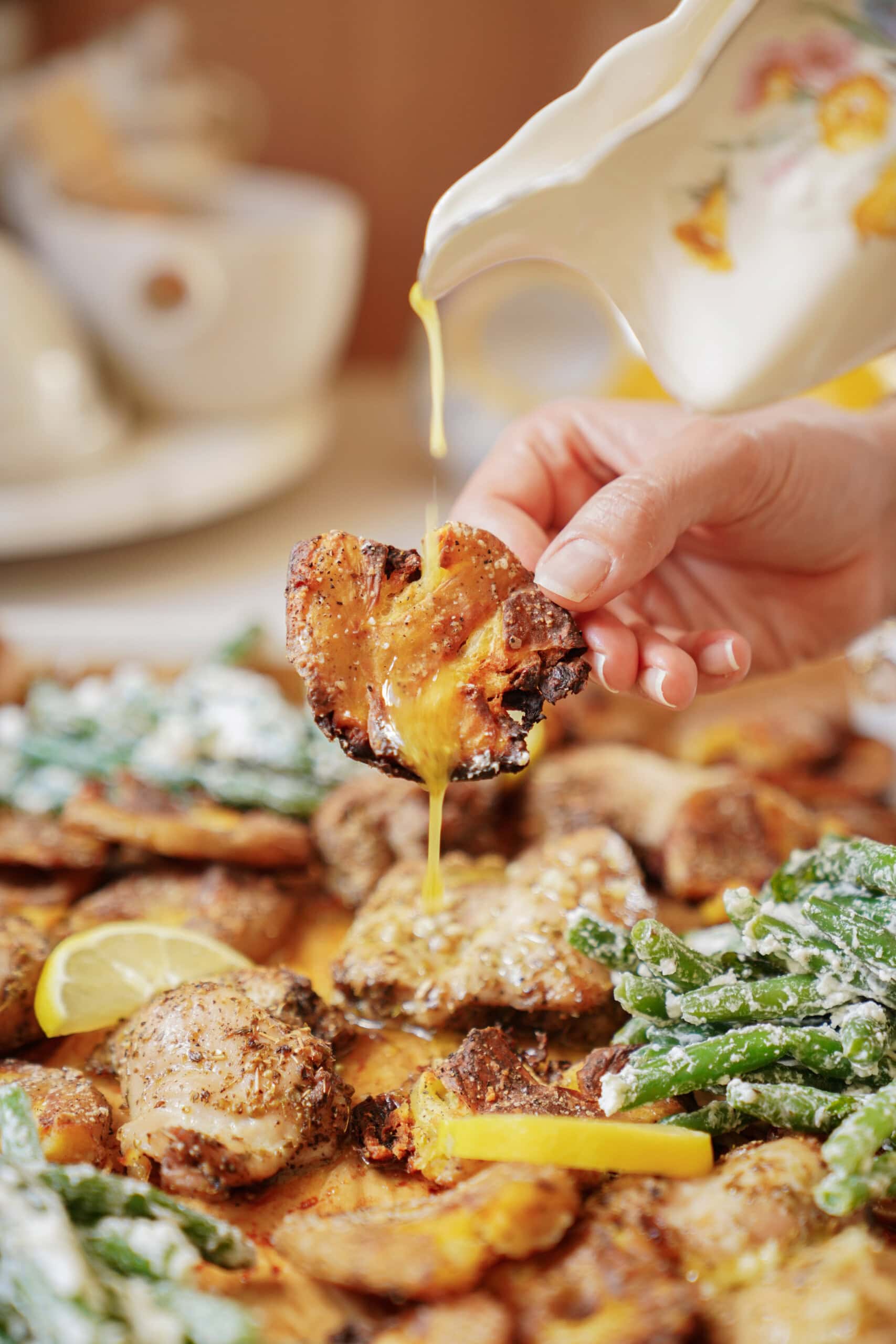 A hand holds a piece of roasted potato while yellow sauce is being poured over it. The potato is surrounded by vegetables, pieces of chicken, and lemon wedges on a serving platter.