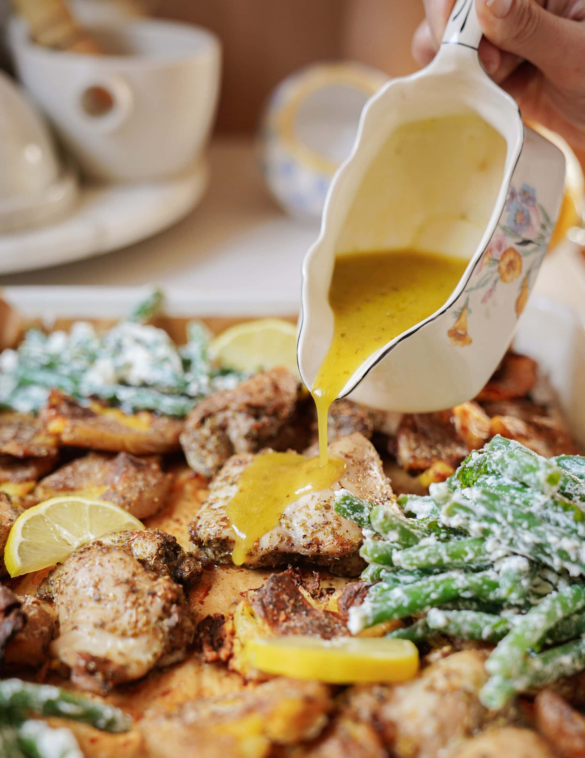 A hand pours yellow sauce from a floral-patterned gravy boat onto seasoned chicken, green beans, and roasted potatoes garnished with lemon slices on a serving tray.