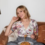 Monique Volz smiling woman with blonde hair wearing a floral blouse and plaid pants sits on a couch, holding a forkful of food above a plate of pasta on her lap.