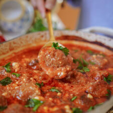 A close-up of a spoon holding a meatball over a pot filled with meatballs in tomato sauce, garnished with parsley.