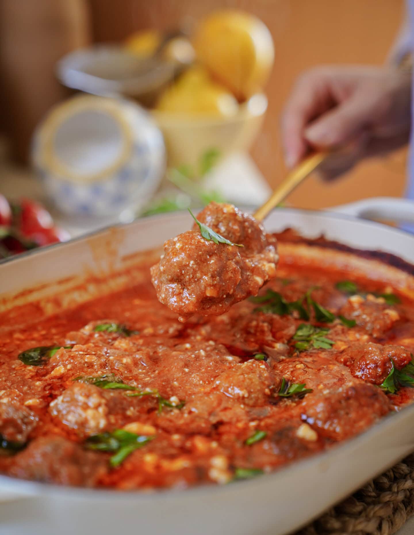 A hand holds a spoon lifting meatballs covered in tomato sauce from a baking dish, garnished with fresh basil. In the background, bowls and lemons are slightly out of focus.