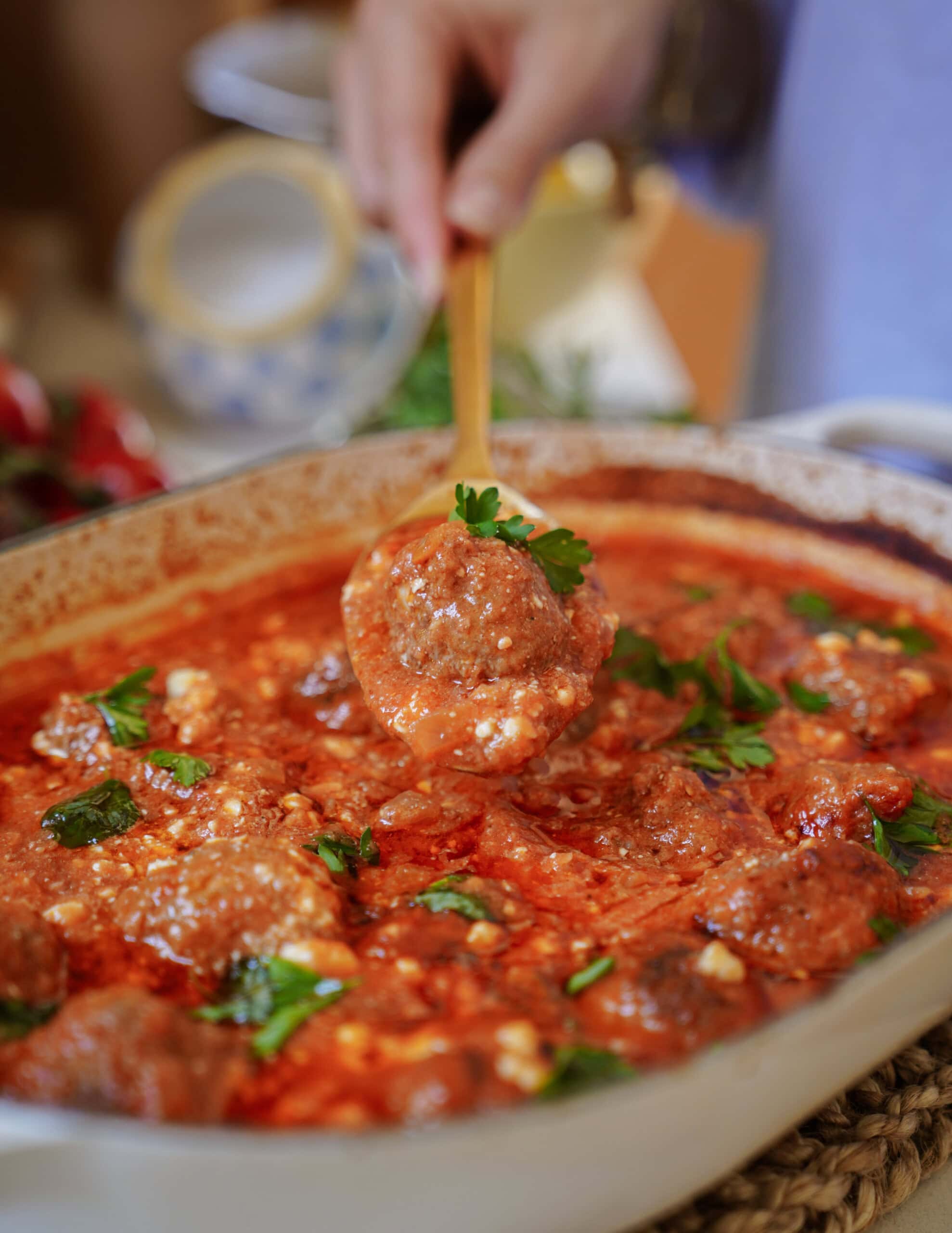 A hand holds a spoon lifting a meatball from a dish filled with meatballs in tomato sauce, garnished with fresh parsley.