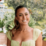 Rachael DeVaux with long brown hair tied back, wearing a light yellow, pleated dress and large hoop earrings, smiles outdoors with lush green plants and trees in the background.