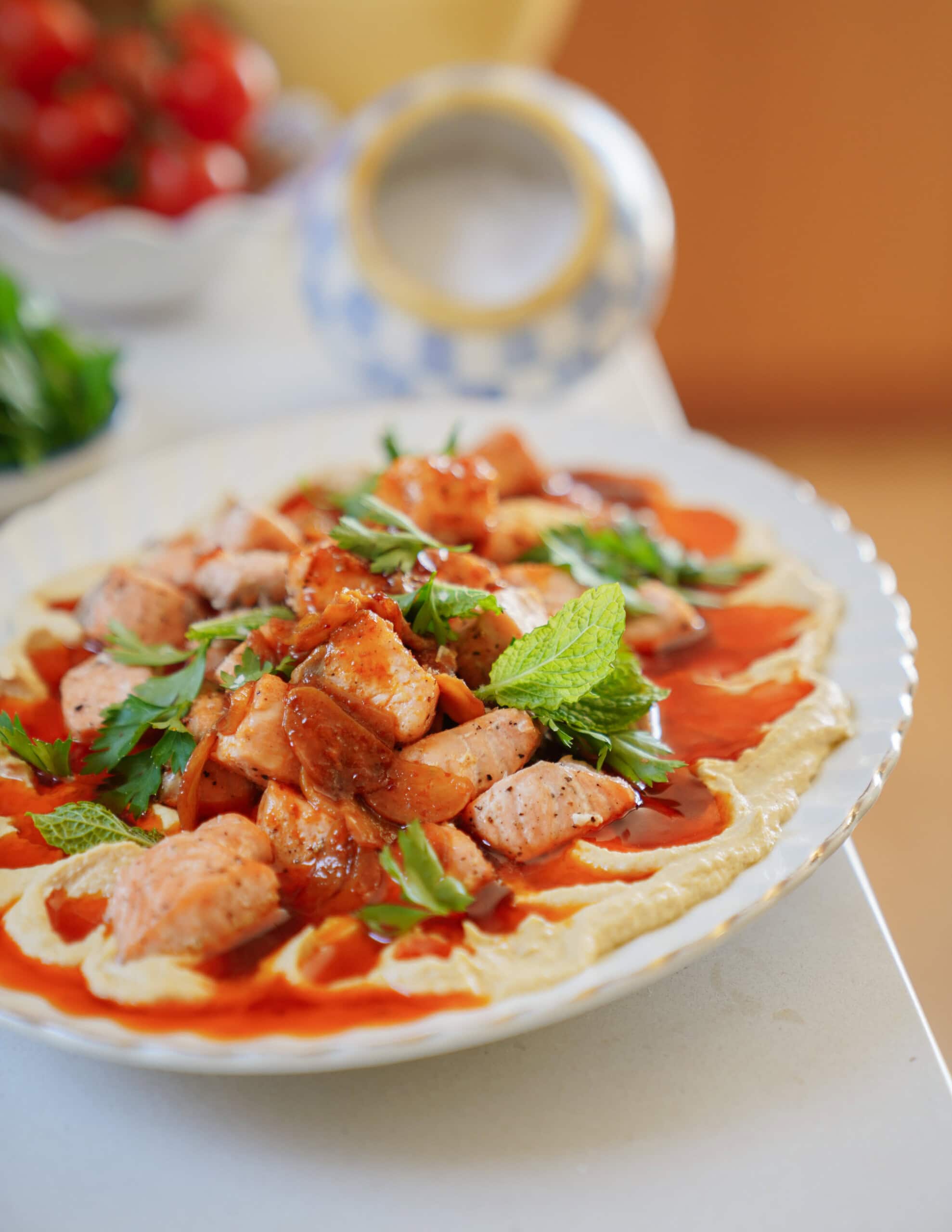 A plate of grilled salmon pieces served on a bed of creamy hummus, topped with fresh parsley and mint leaves, drizzled with a red sauce, with a blurred bowl and tomatoes in the background.
