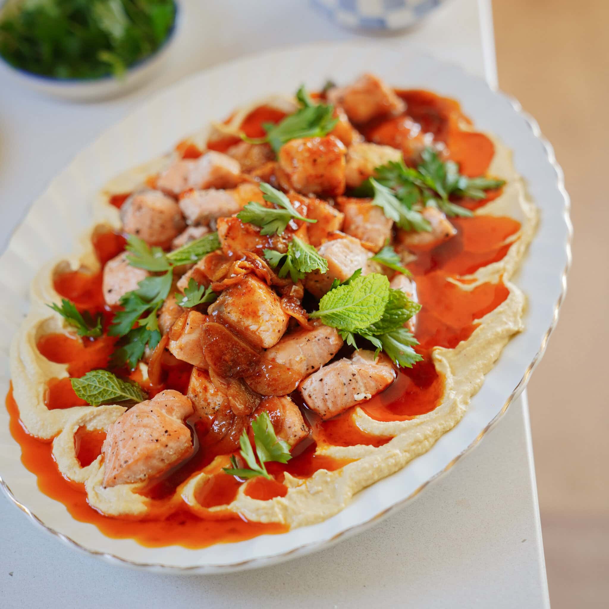 A white platter with creamy hummus topped with chunks of grilled chicken, drizzled with red sauce, and garnished with fresh parsley and mint leaves. The dish is set on a white countertop.