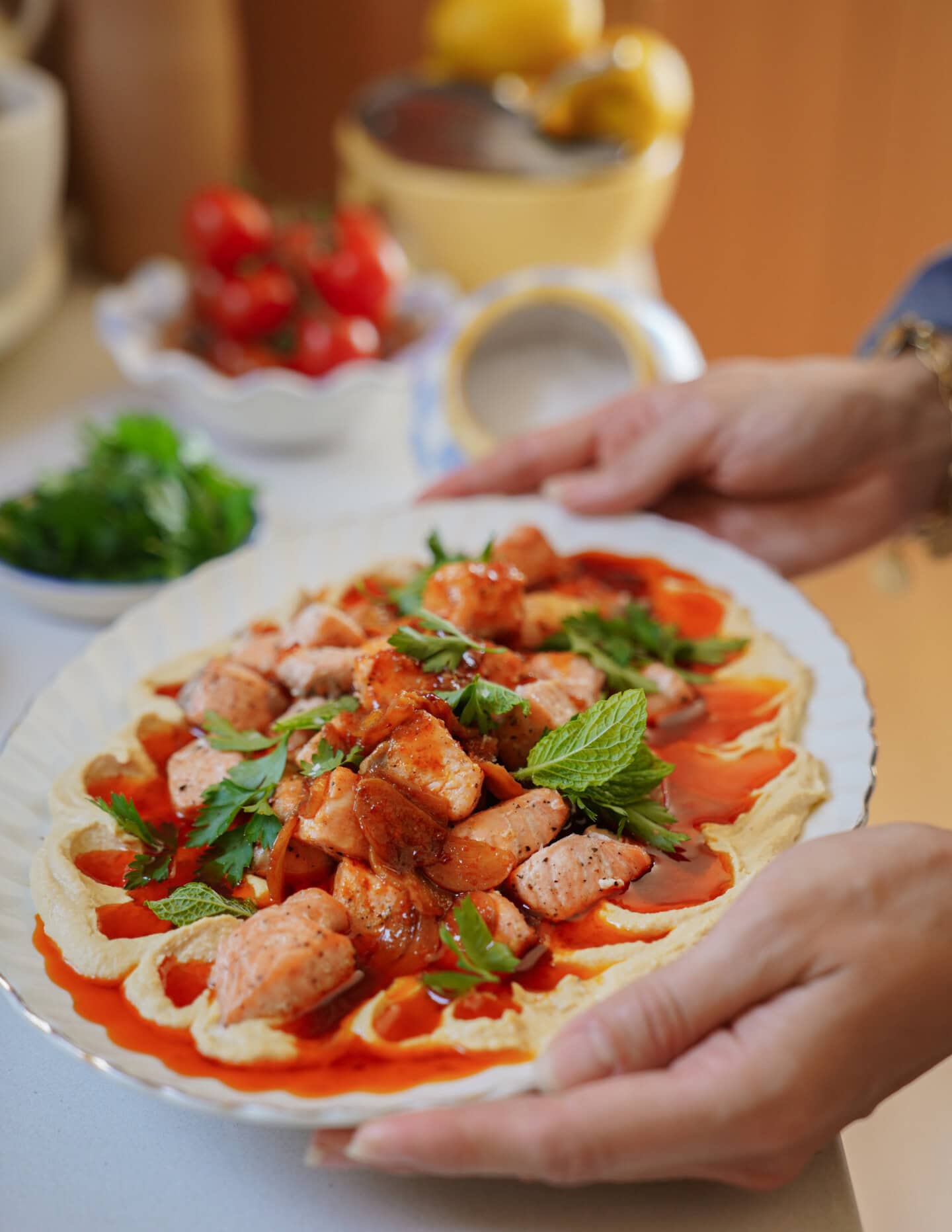 Hands holding a plate of food topped with pieces of grilled salmon, fresh herbs, and a red sauce, with ingredients like tomatoes and greens blurred in the background.