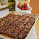 A baking tray with rows of cooked minced meat, possibly kebab or meatloaf, sits on a kitchen counter. In the background, there is a bottle, a bowl, and a dish of cherry tomatoes.