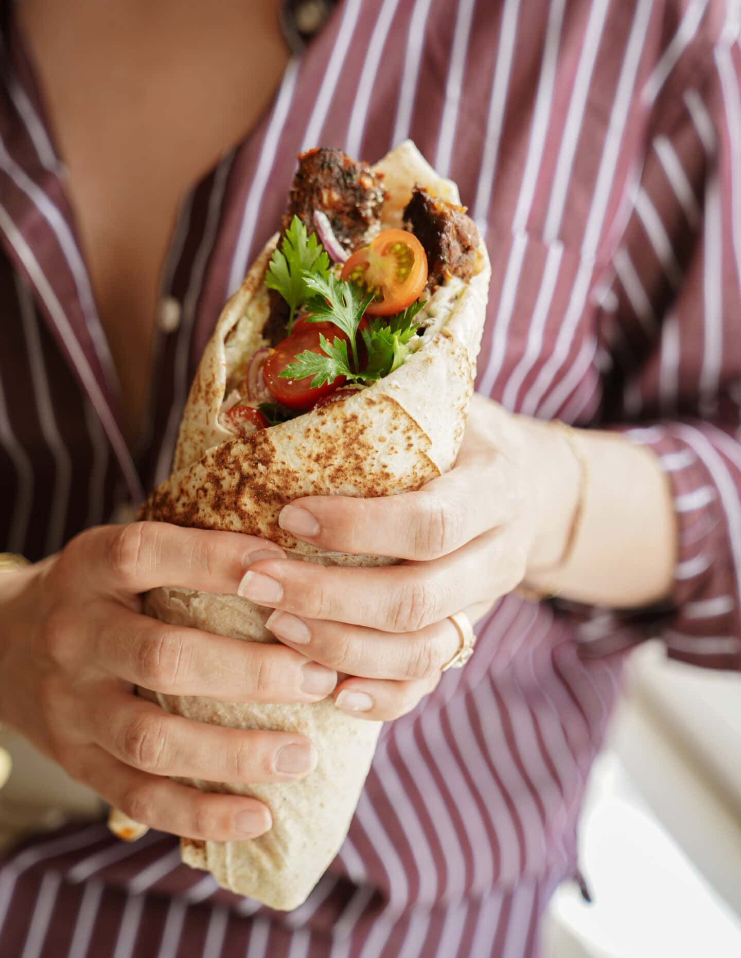 A person in a maroon and white striped shirt holds a large wrap filled with grilled meat, cherry tomatoes, and fresh cilantro.