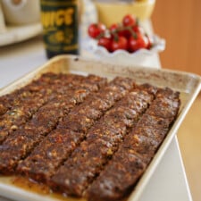 A baking tray with rows of cooked minced meat, possibly kebab or meatloaf, sits on a kitchen counter. In the background, there is a bottle, a bowl, and a dish of cherry tomatoes.