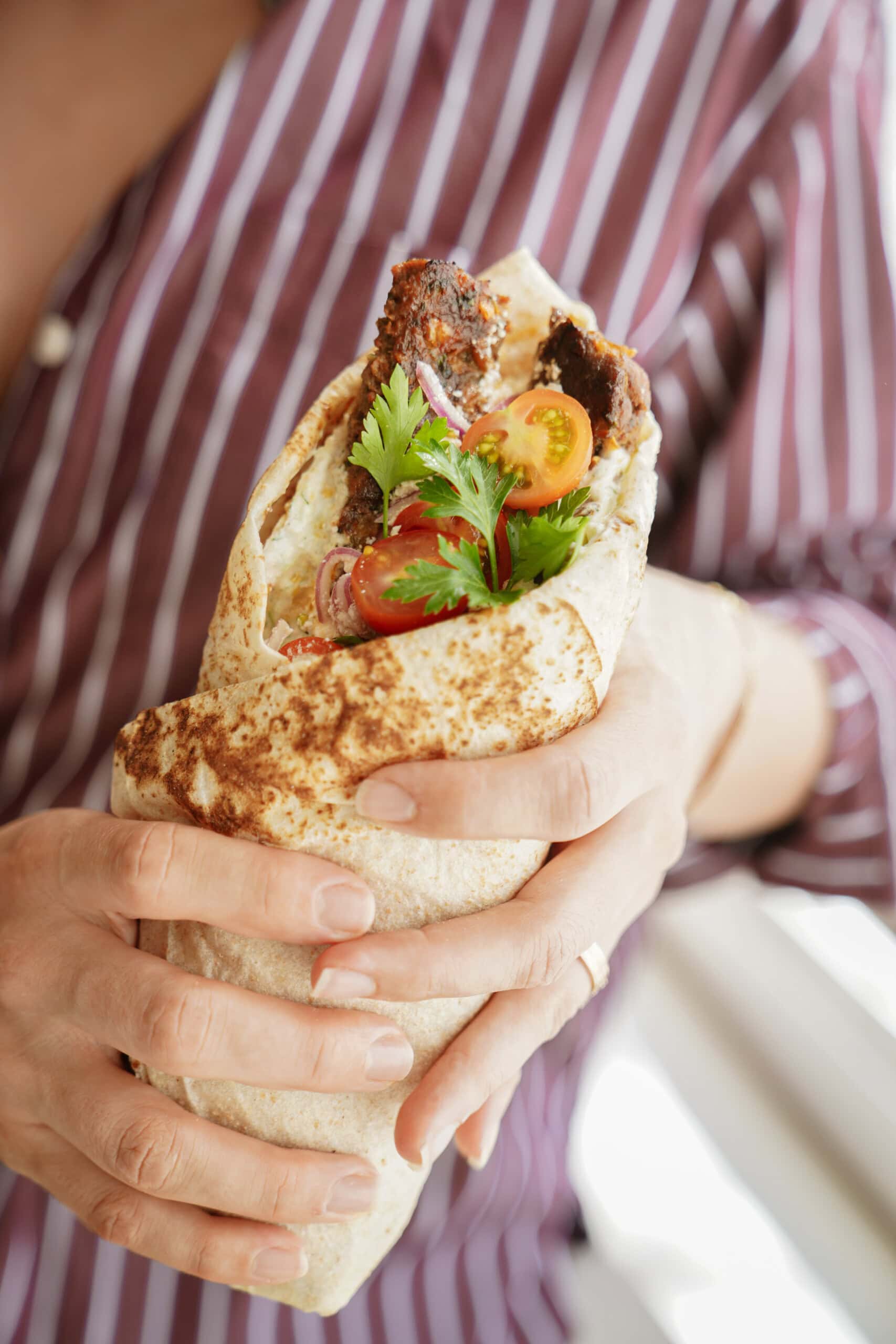 A person in a maroon and white striped shirt holds a large wrap filled with grilled meat, cherry tomatoes, red onions, and fresh cilantro.