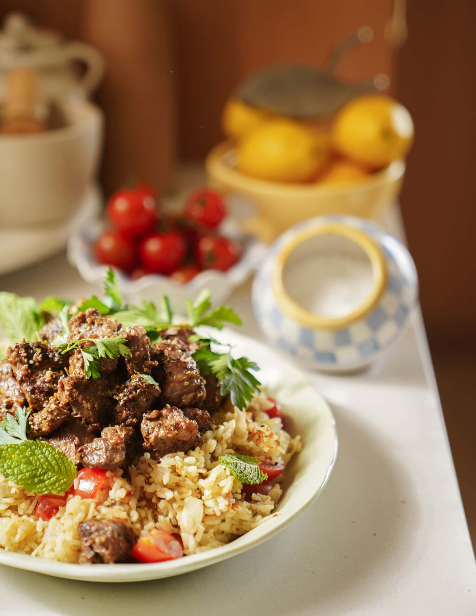 A plate of seasoned rice with chopped tomatoes, topped with spiced meat and fresh herbs, sits on a kitchen counter. In the background are a bowl of cherry tomatoes, lemons, and a decorative salt cellar.