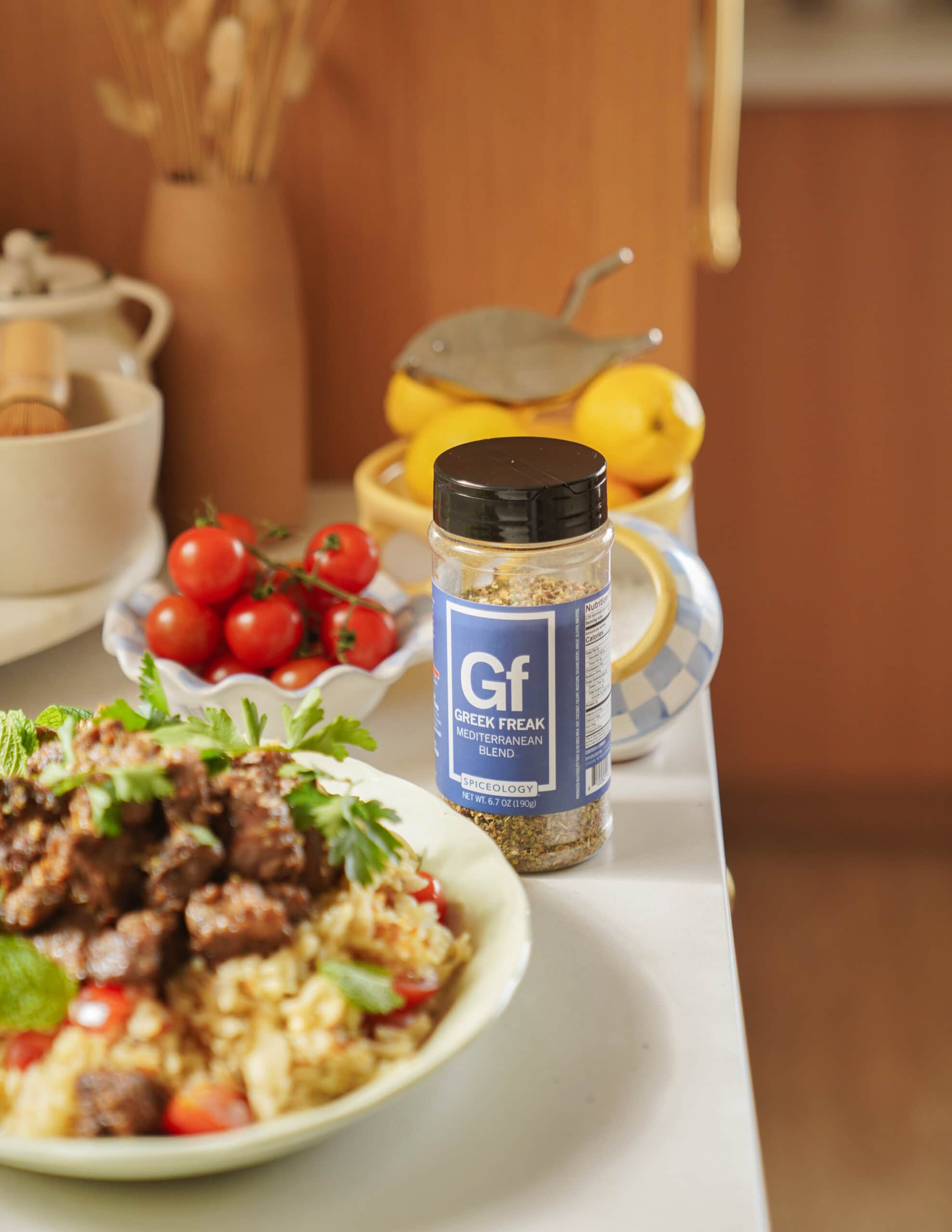 A jar labeled Gf Greek Freak Mediterranean seasoning sits on a kitchen counter near a bowl of food with meat and rice, fresh tomatoes, lemons, and kitchen items in the background.