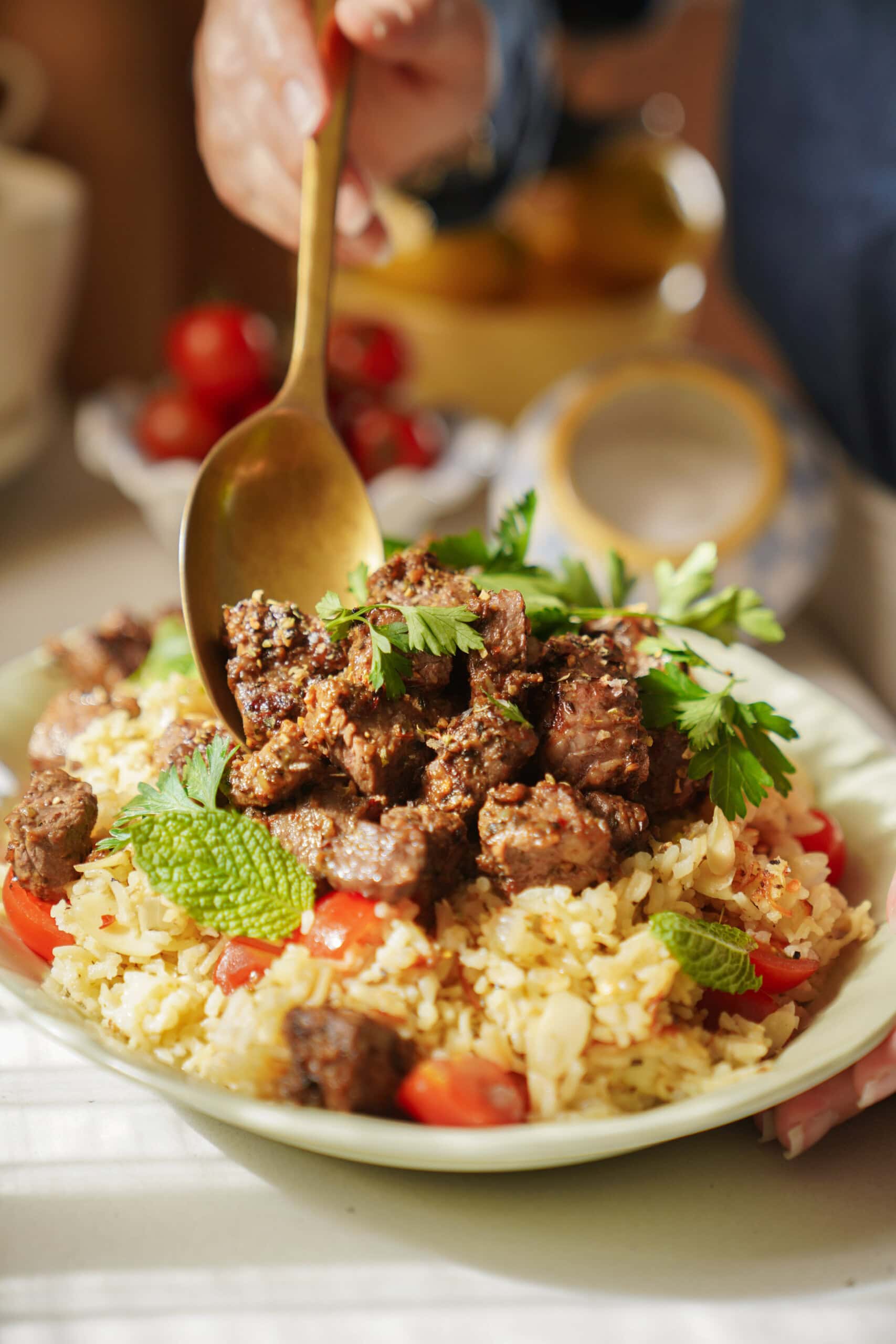 A hand serves spiced, grilled meat pieces over a plate of rice garnished with fresh parsley, mint, almond slivers, and tomato slices.