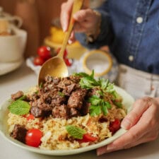 A person serves seasoned grilled beef cubes onto a plate of rice with cherry tomatoes, garnished with fresh mint and parsley.