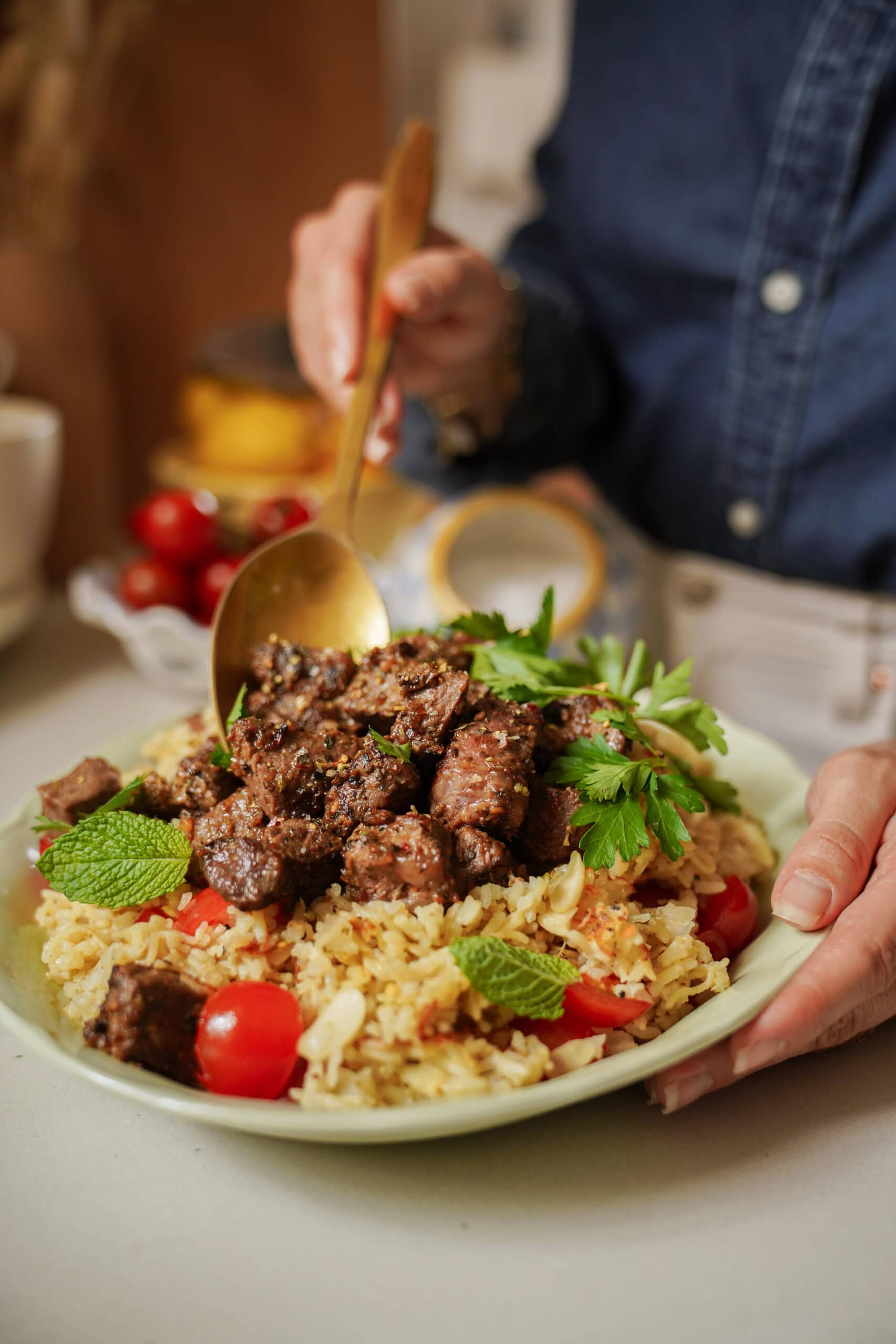 A person in a blue shirt serves seasoned grilled meat over a plate of rice with cherry tomatoes and fresh herbs, using a gold spoon.