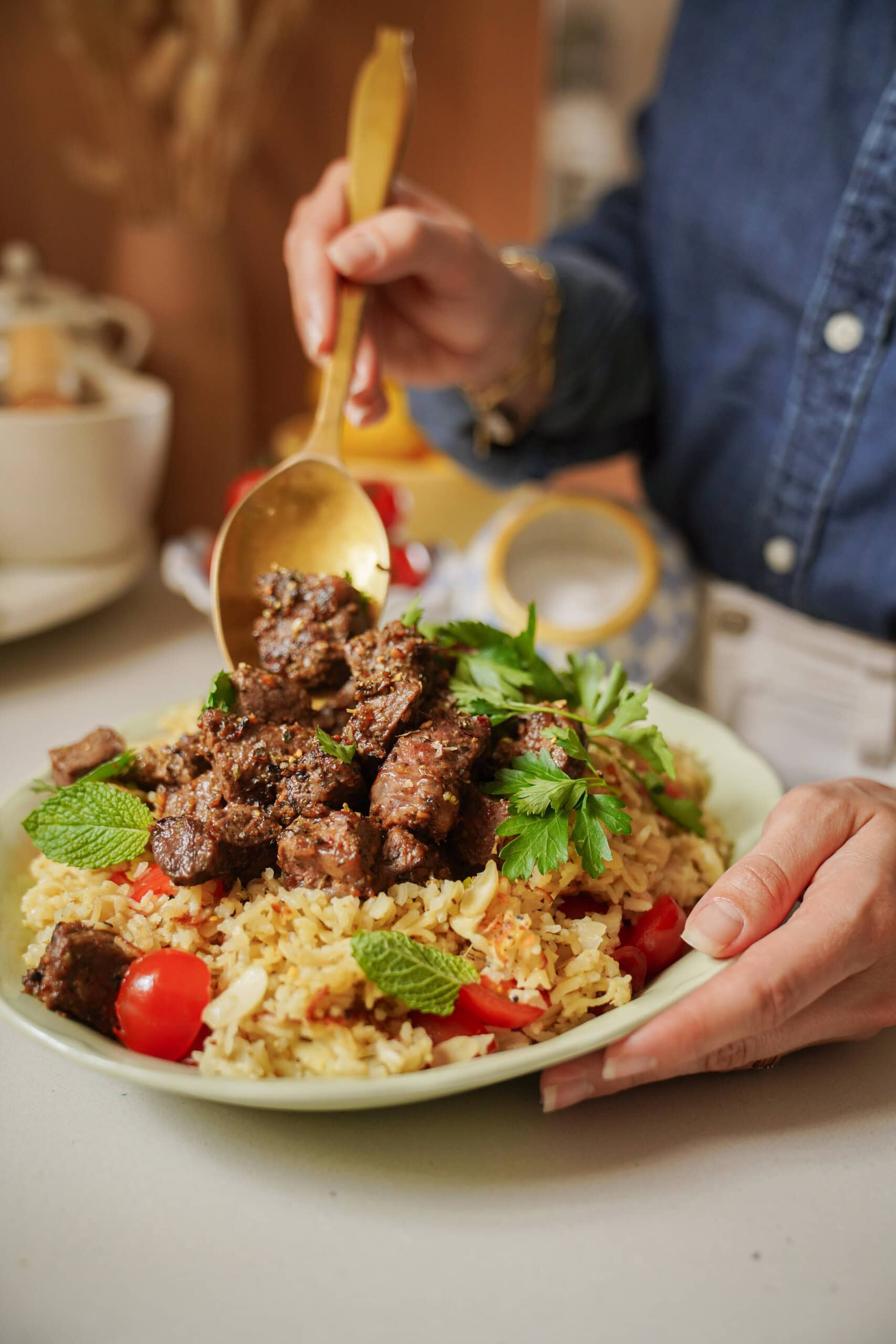 A person in a blue shirt serves seasoned beef chunks onto a plate of rice, garnished with fresh herbs and cherry tomatoes, using a large golden spoon.