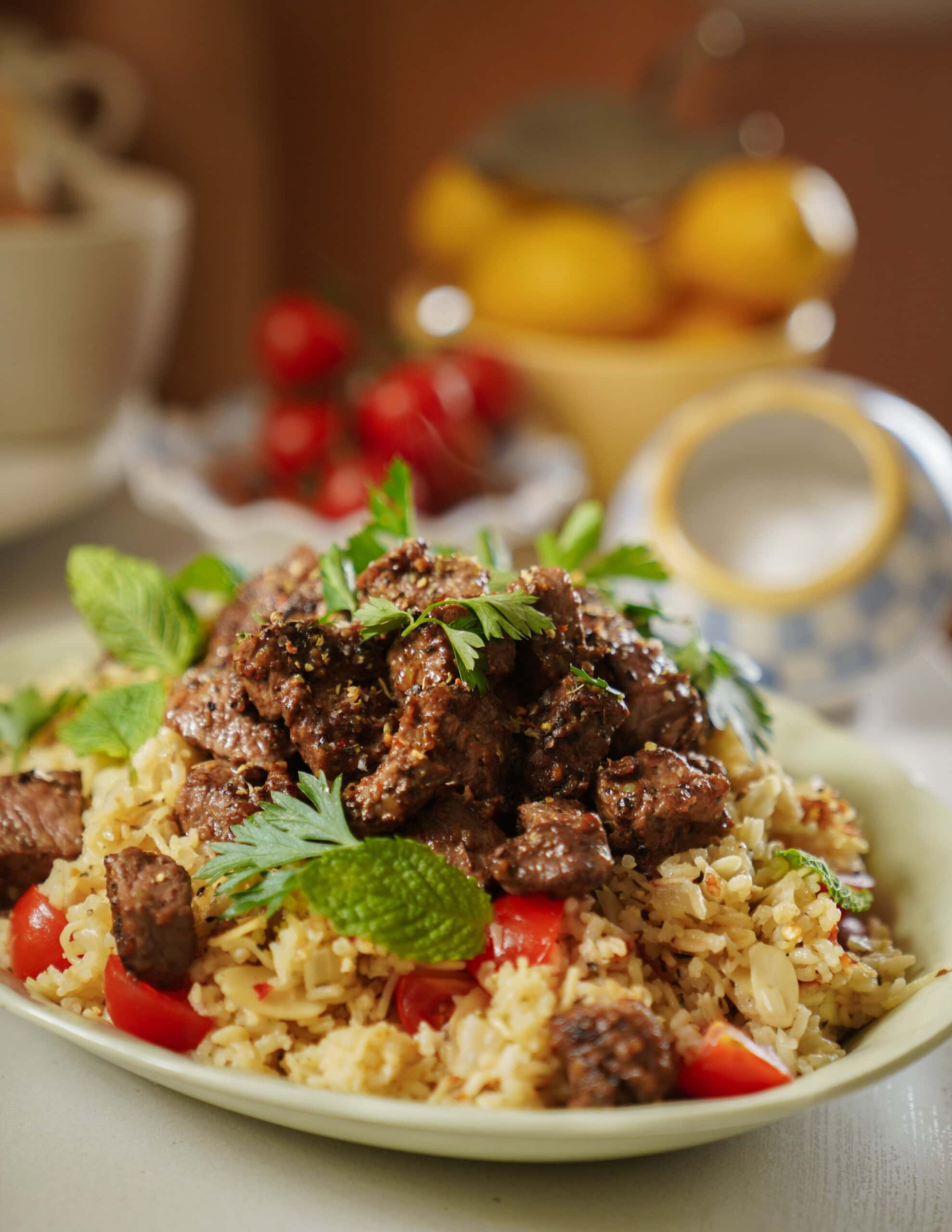 A plate of rice pilaf topped with seasoned beef cubes, garnished with fresh mint and parsley, with cherry tomatoes mixed in. Blurred background includes lemons and a teapot.