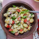 A wooden bowl filled with potato salad containing halved cherry tomatoes, red onion, crumbled cheese, capers, and fresh basil leaves, with a spoon resting inside.