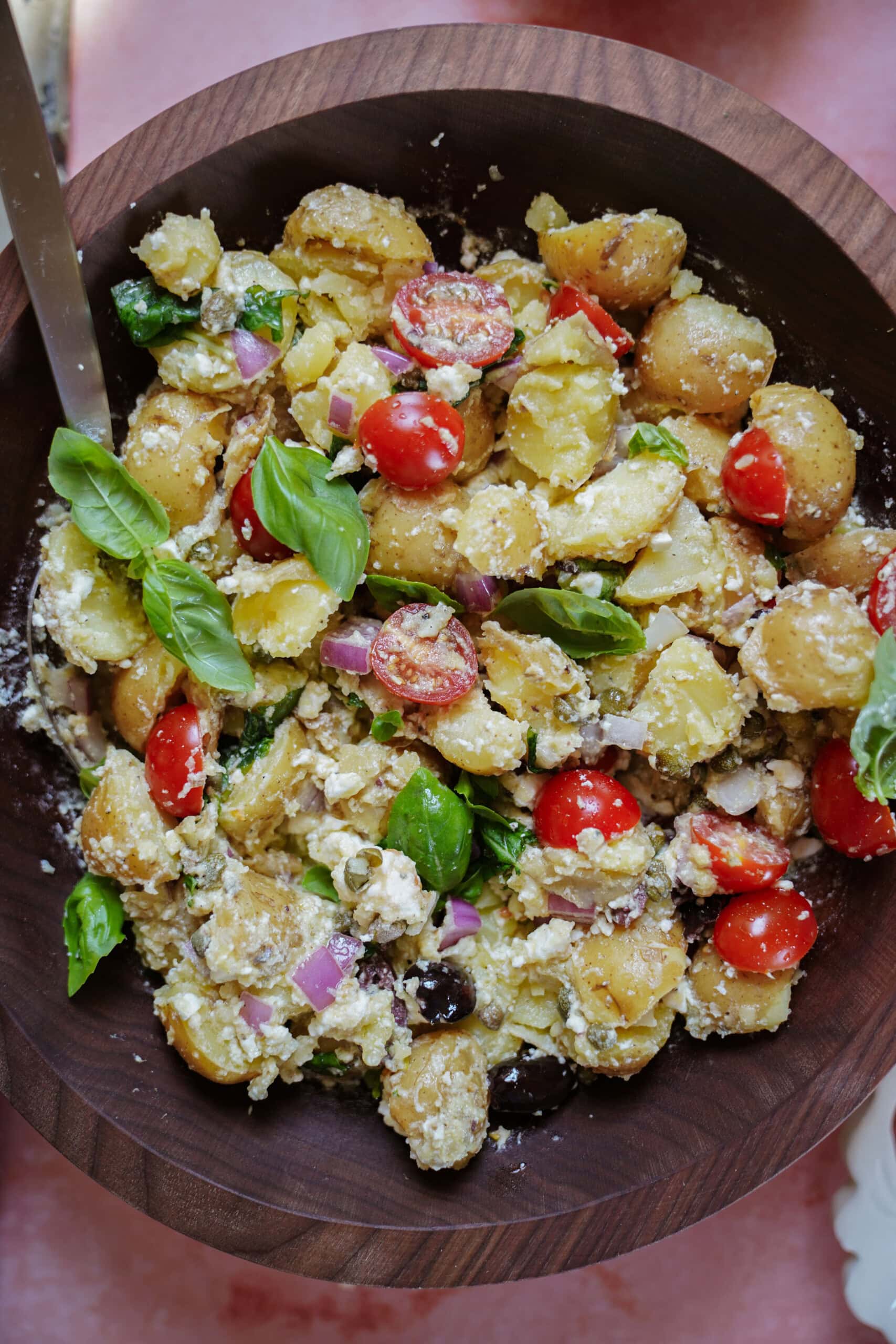A wooden bowl filled with potato salad, featuring halved cherry tomatoes, red onion, fresh basil leaves, crumbled cheese, and black olives. A metal serving spoon rests on the side of the bowl.