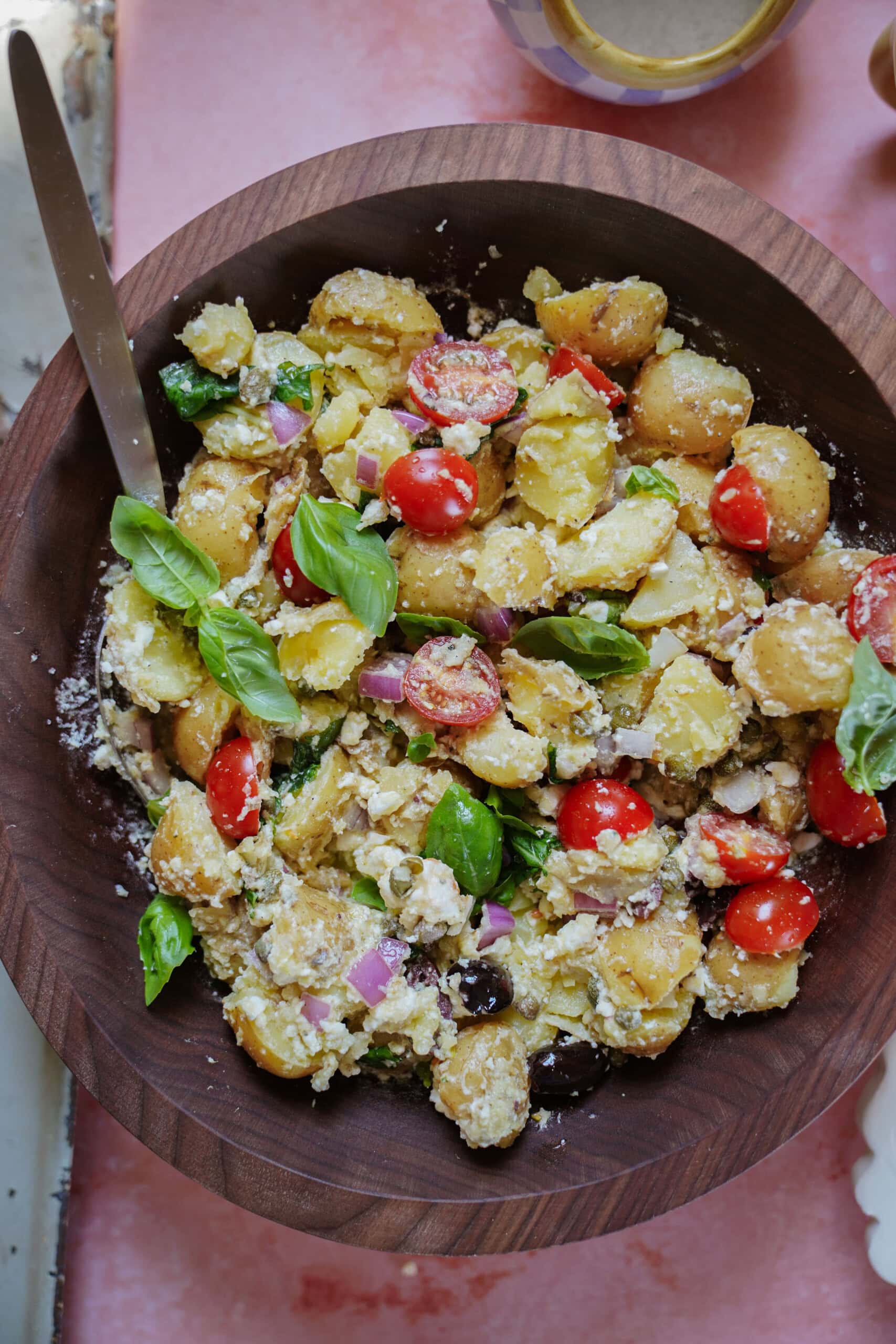 A wooden bowl filled with a colorful potato salad, featuring cherry tomatoes, red onion, olives, fresh basil leaves, and crumbled cheese, all mixed together.