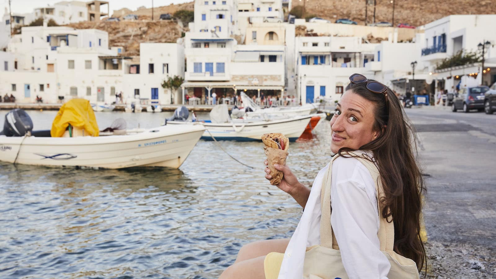A woman with long brown hair sits by the waterfront, holding an ice cream cone and smiling at the camera. Behind her are boats docked along the water and white buildings with blue accents.