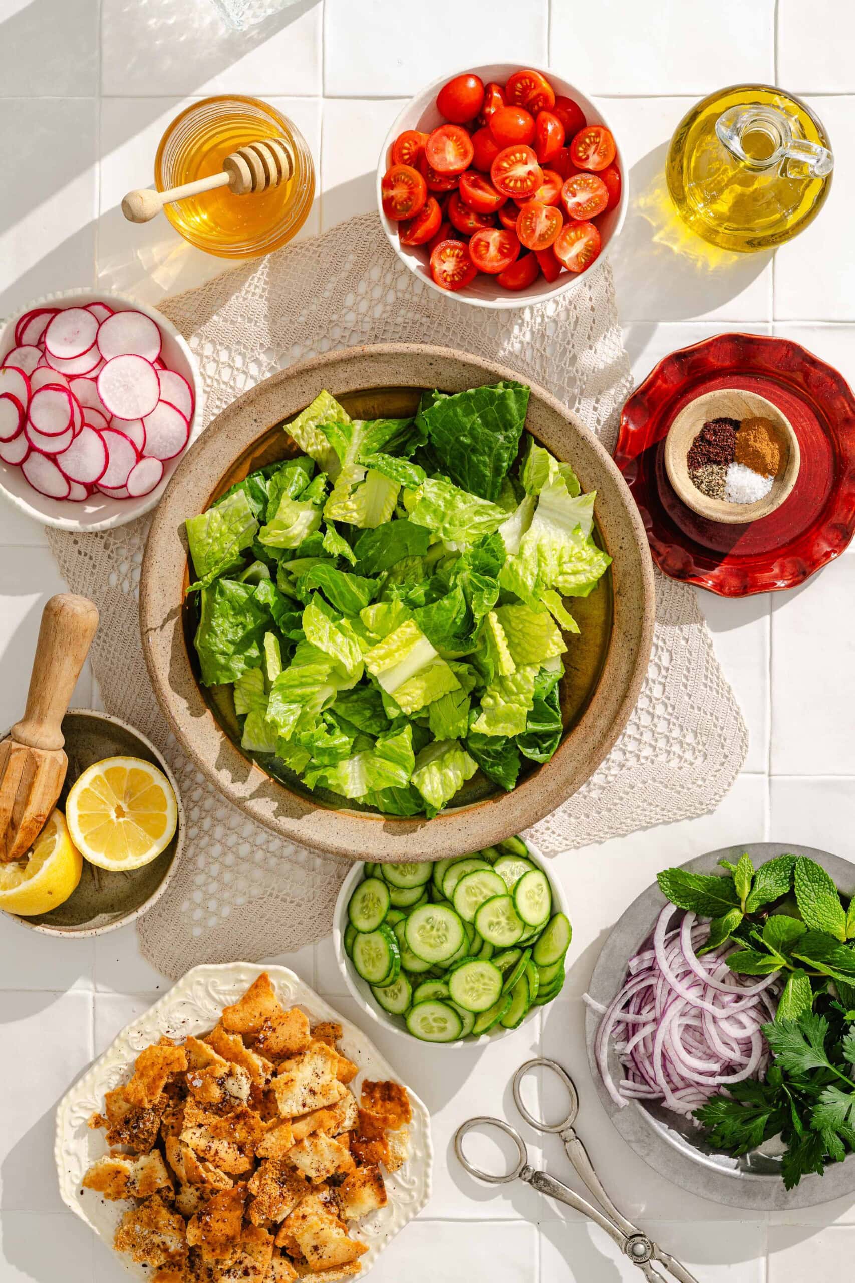 A top-down view of a salad-making setup with a bowl of chopped lettuce in the center, surrounded by sliced radishes, cherry tomatoes, cucumber, red onion, herbs, croutons, lemon, a jar of honey, olive oil, and seasonings.