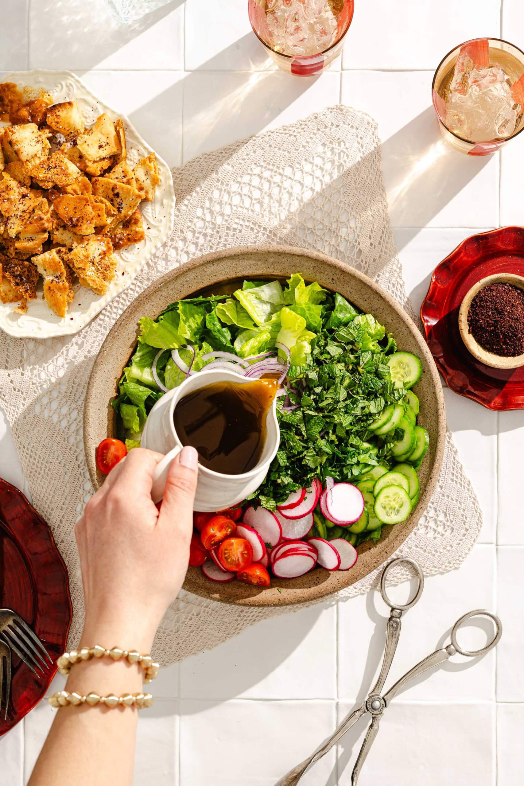 A hand pours dark dressing over a fresh salad with lettuce, cucumber, radish, tomato, and herbs in a bowl. Nearby are croutons, a bowl of spices, scissors, and glasses of ice on a bright table.
