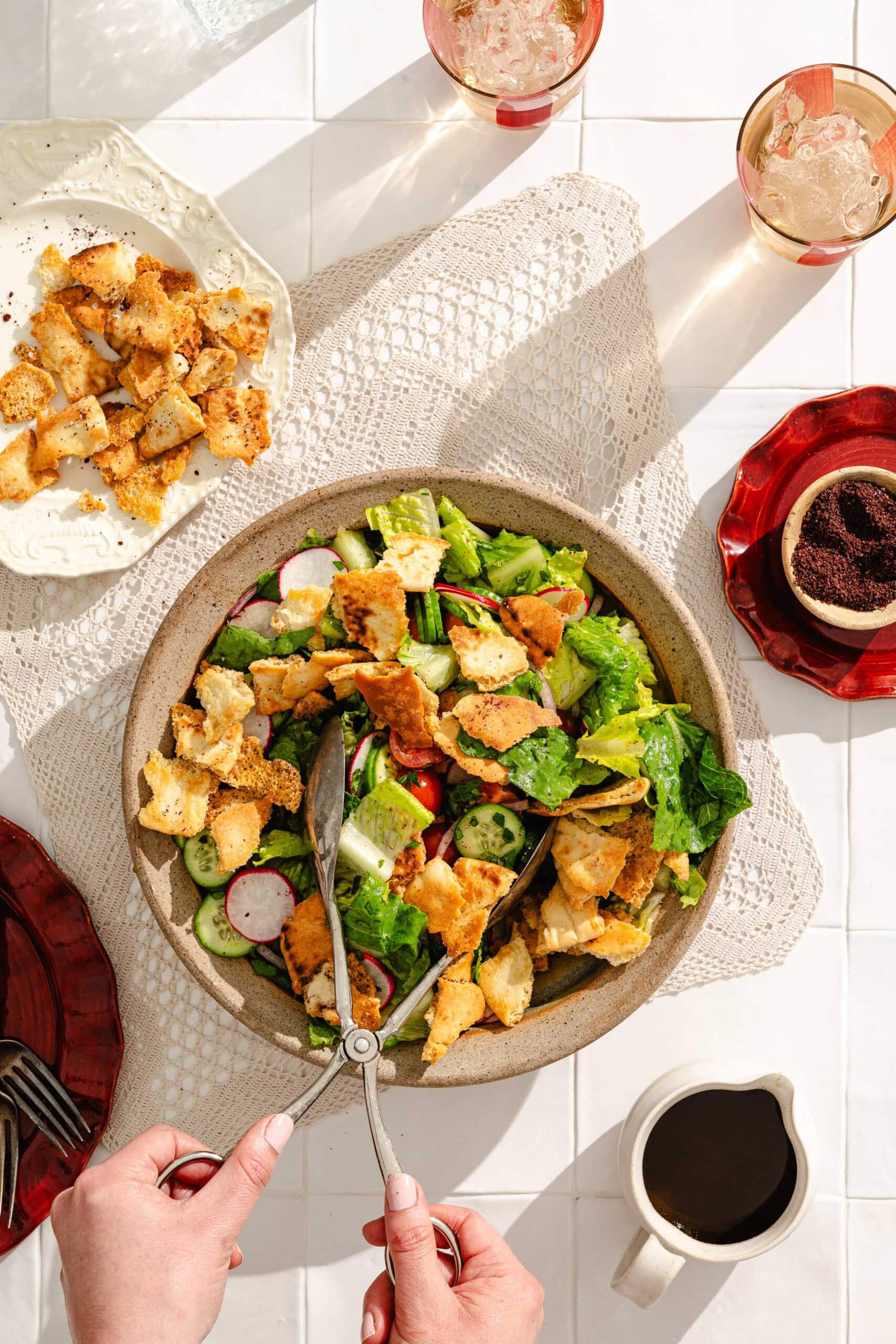 A person uses tongs to serve a fresh salad with lettuce, radishes, cucumbers, tomatoes, and crispy pita chips. Surrounding items include cut pita, drinks, coffee, and a lace table runner on a white tile surface.
