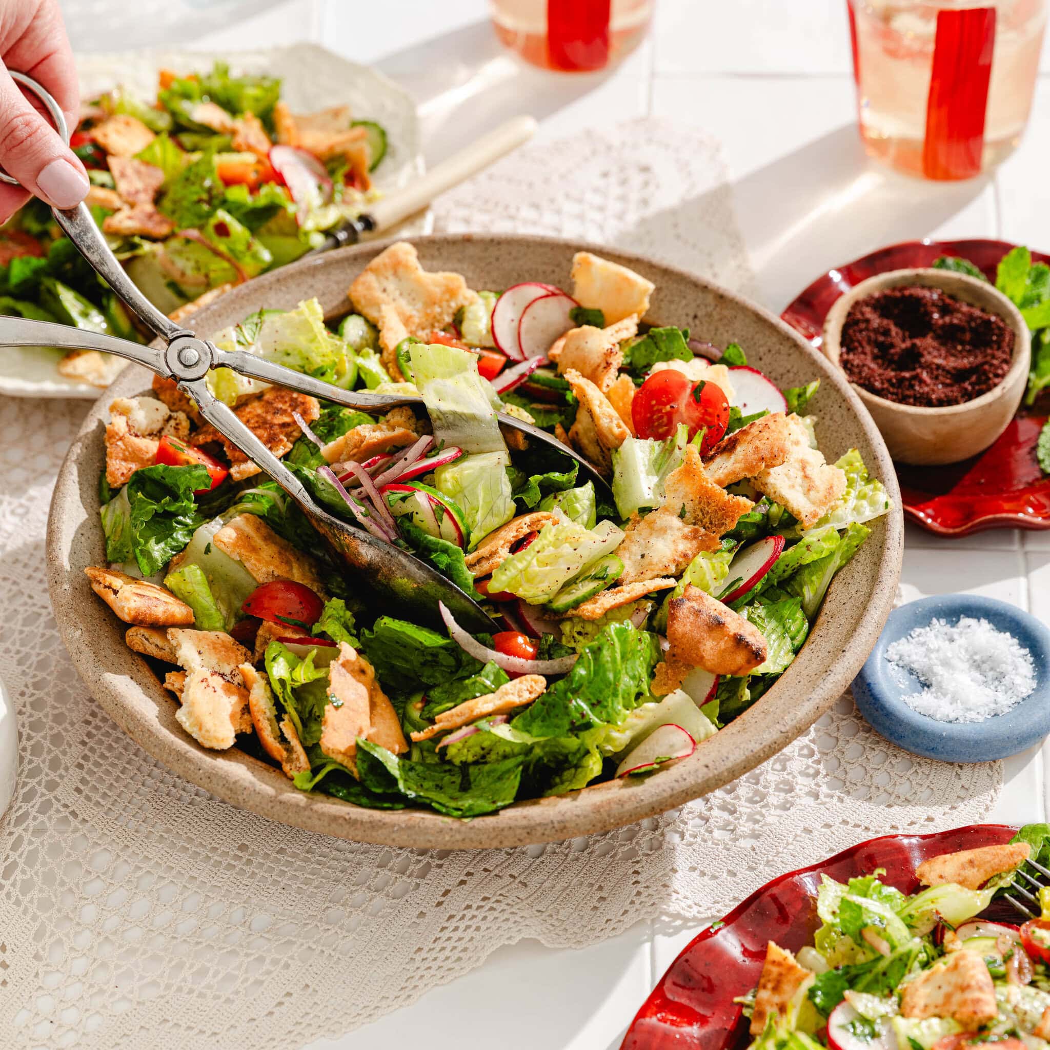 A hand uses tongs to serve a fresh salad with lettuce, radish, tomatoes, and crispy pita pieces from a large bowl, surrounded by red plates, drinks, and small bowls of spices on a white lace tablecloth.