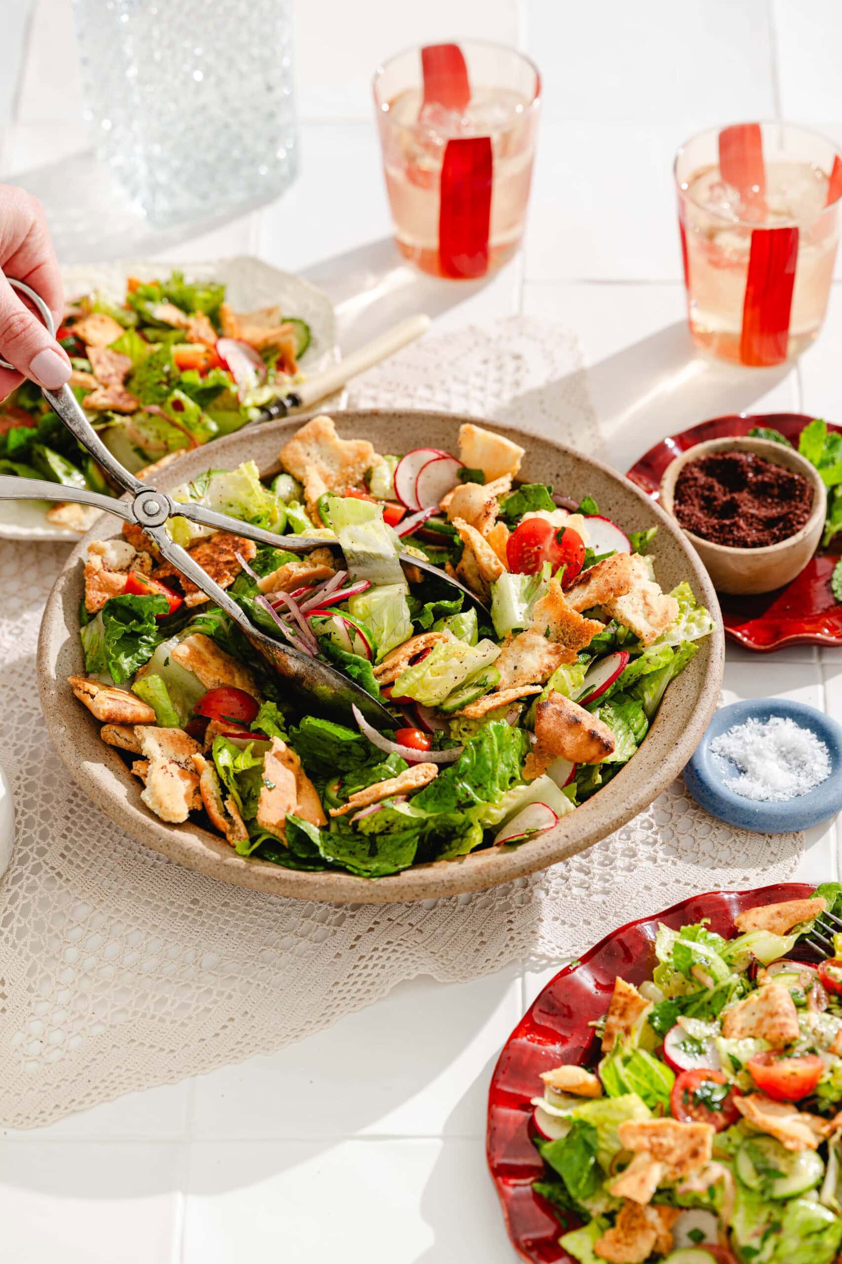 A hand tosses a fresh salad with tongs in a large bowl, surrounded by plates of salad, drinks in glasses with red stripes, and small dishes of salt and spices on a sunlit table.