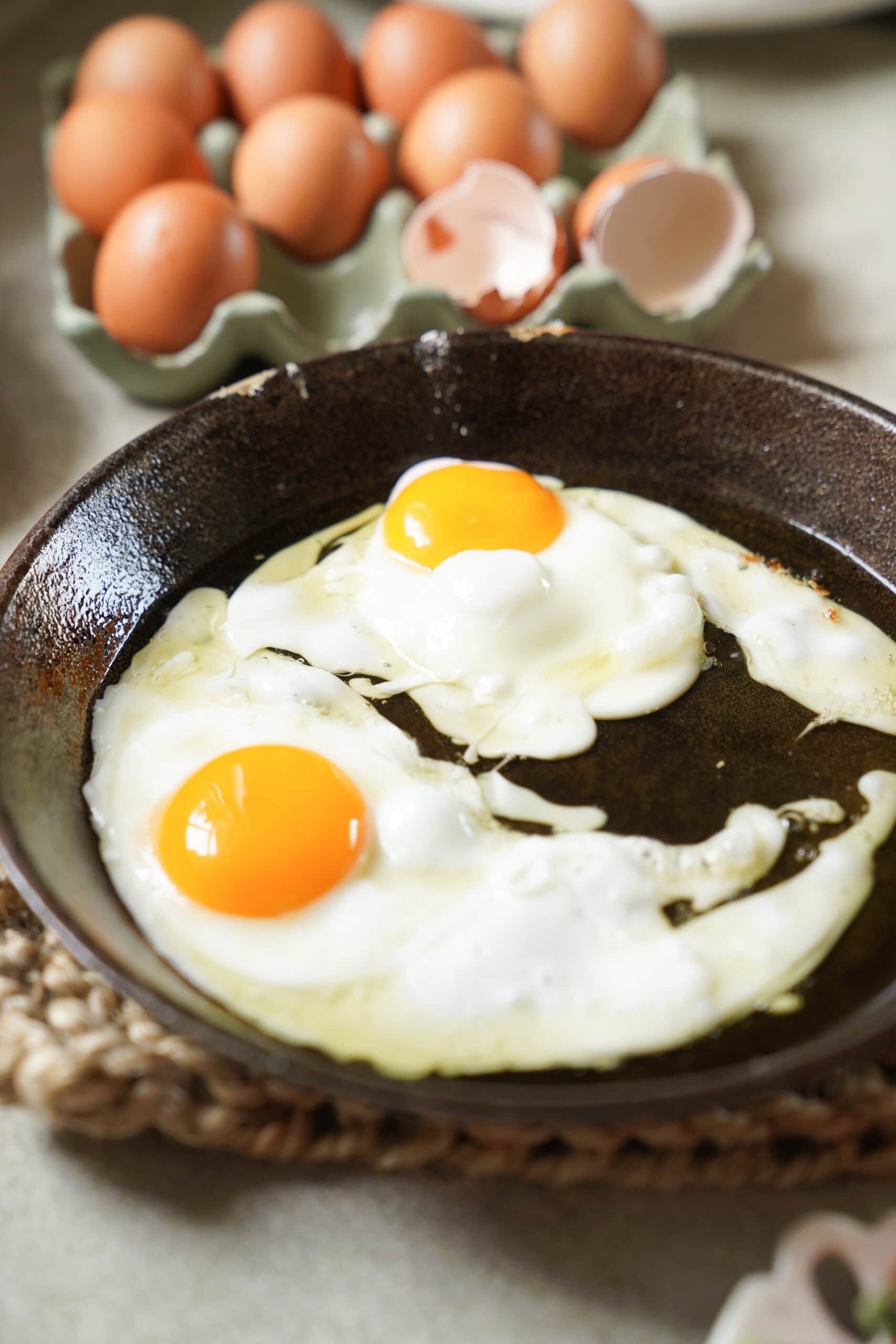Two sunny-side-up eggs cooking in a dark skillet, with a carton of brown eggs and some cracked eggshells in the background.