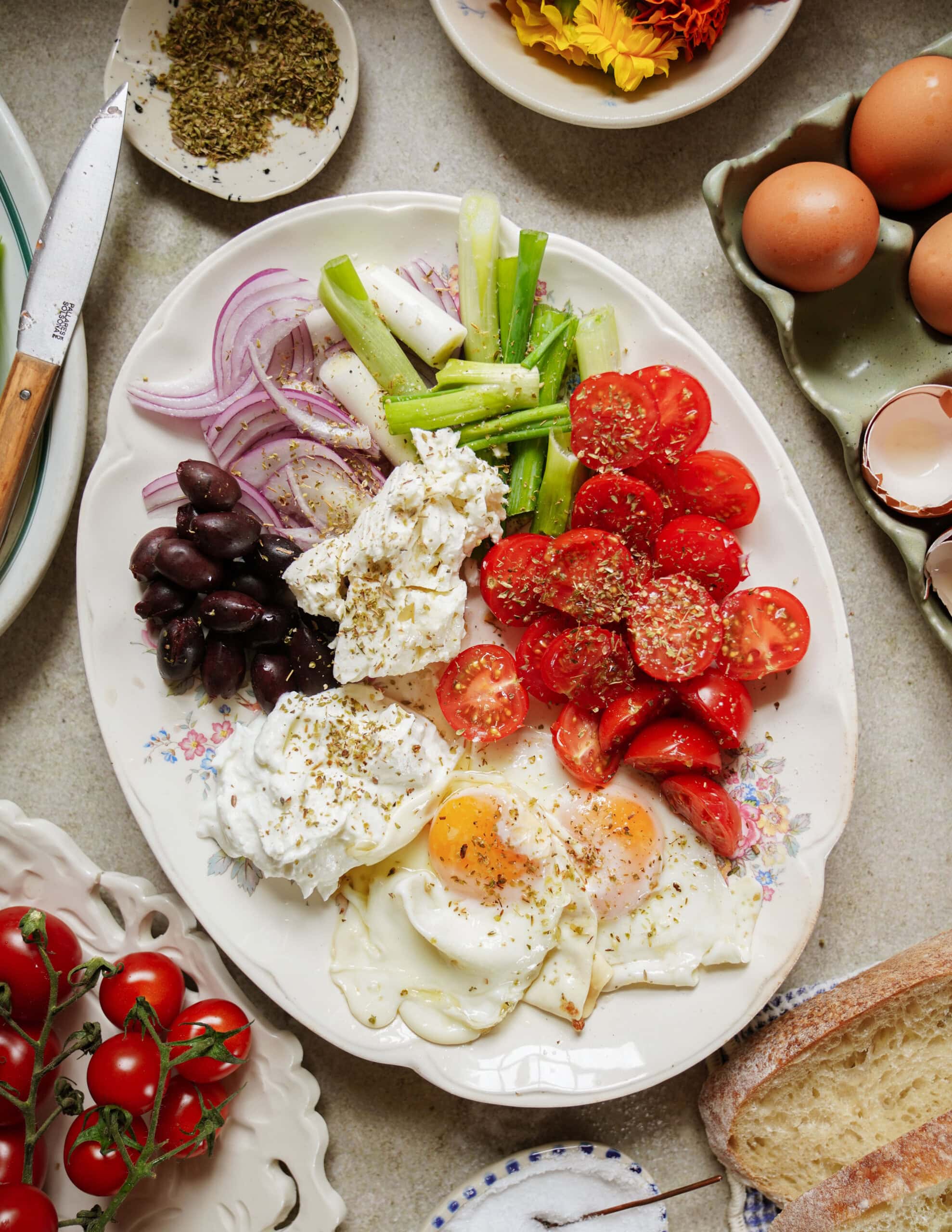 A plate with fried eggs, sliced tomatoes, burrata cheese, black olives, red onion, and green onions, garnished with herbs. Surrounding the plate are eggs, cherry tomatoes, bread, herbs, and flowers.