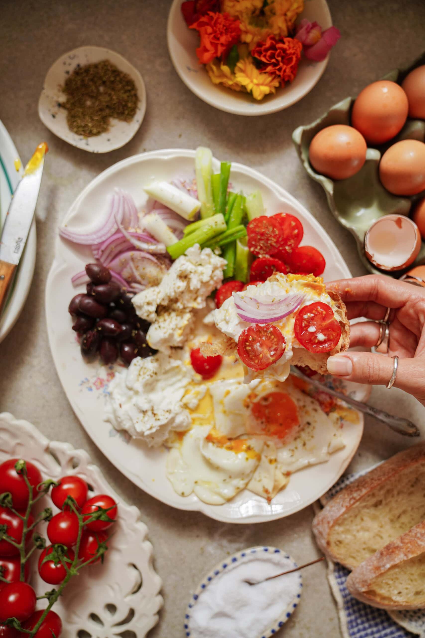 A hand holds toast topped with sliced tomatoes and onions over a platter of fried eggs, tomatoes, olives, cheese, onions, and celery, surrounded by eggs, bread, herbs, and flowers on a table.