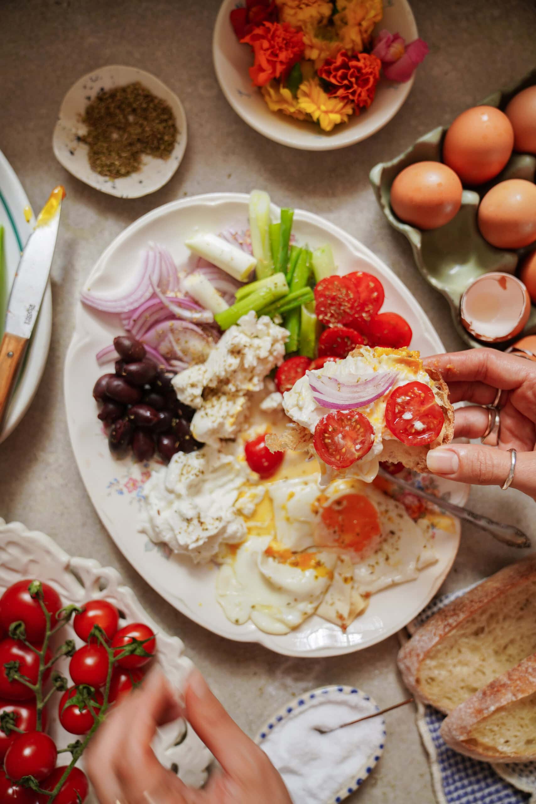A hand holds a slice of bread topped with tomatoes and onions above a platter of sliced onions, celery, olives, cheese, tomatoes, and fried eggs. Surrounding the platter are whole eggs, tomatoes, bread, and a small bowl of salt.