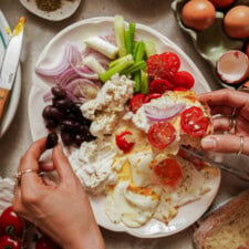 A person prepares toast with fresh tomatoes, red onions, ricotta cheese, and eggs from a platter surrounded by olives, vegetables, whole eggs, and bread.
