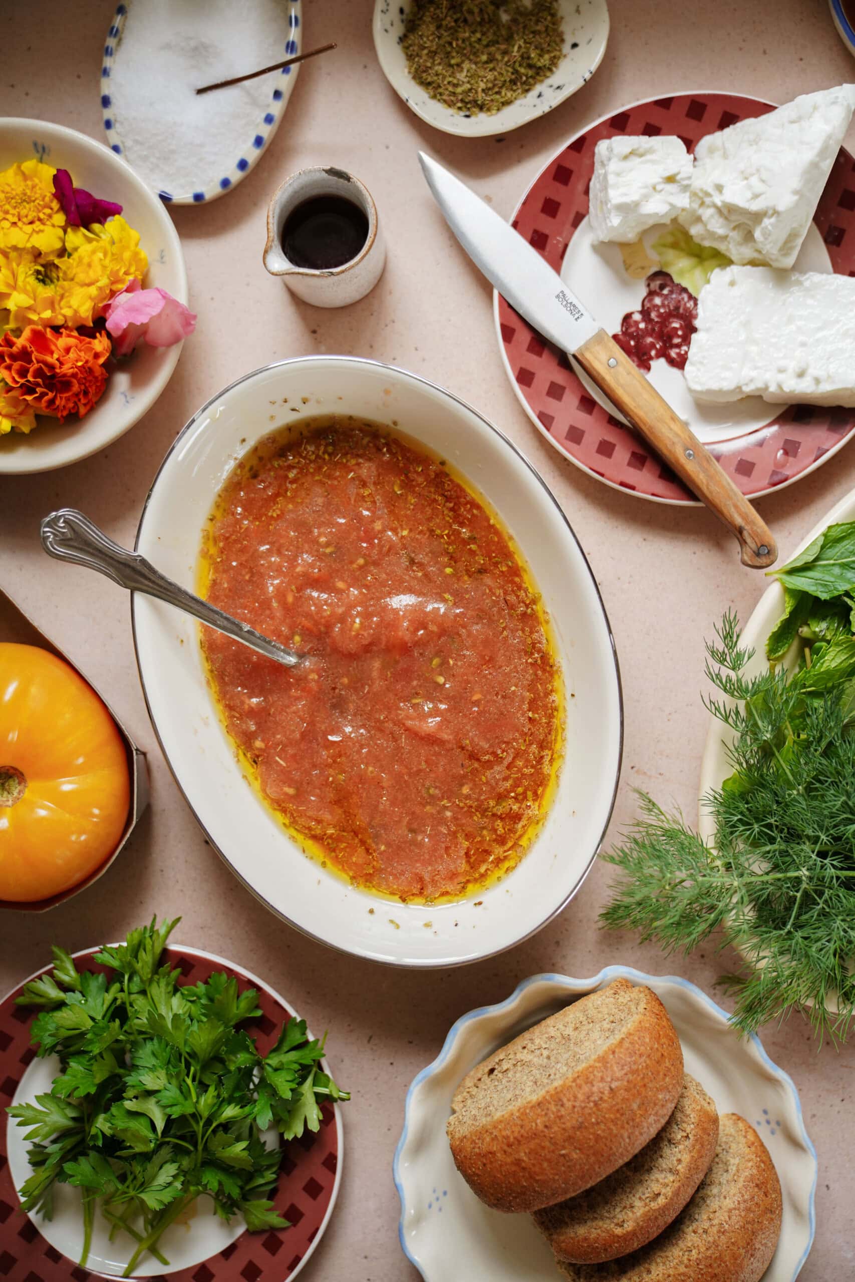 A table set with a bowl of tomato sauce, a spoon, sliced brown bread, fresh herbs, feta cheese, a knife, a yellow tomato, a plate of edible flowers, olive oil, and seasoning.