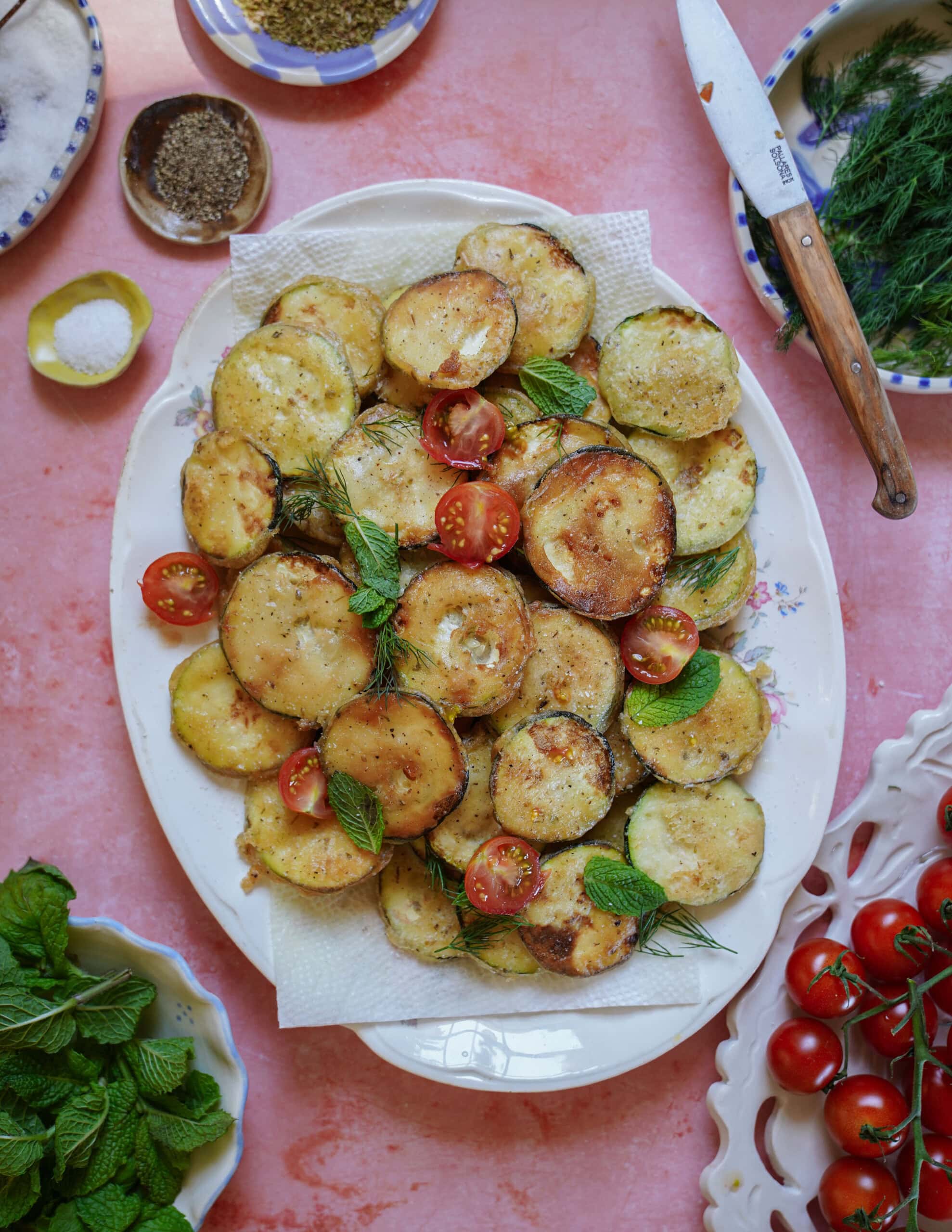 A platter of golden, roasted potato slices garnished with fresh cherry tomato halves, mint leaves, and dill, set on a pink surface with herbs, salt, pepper, and a knife nearby.
