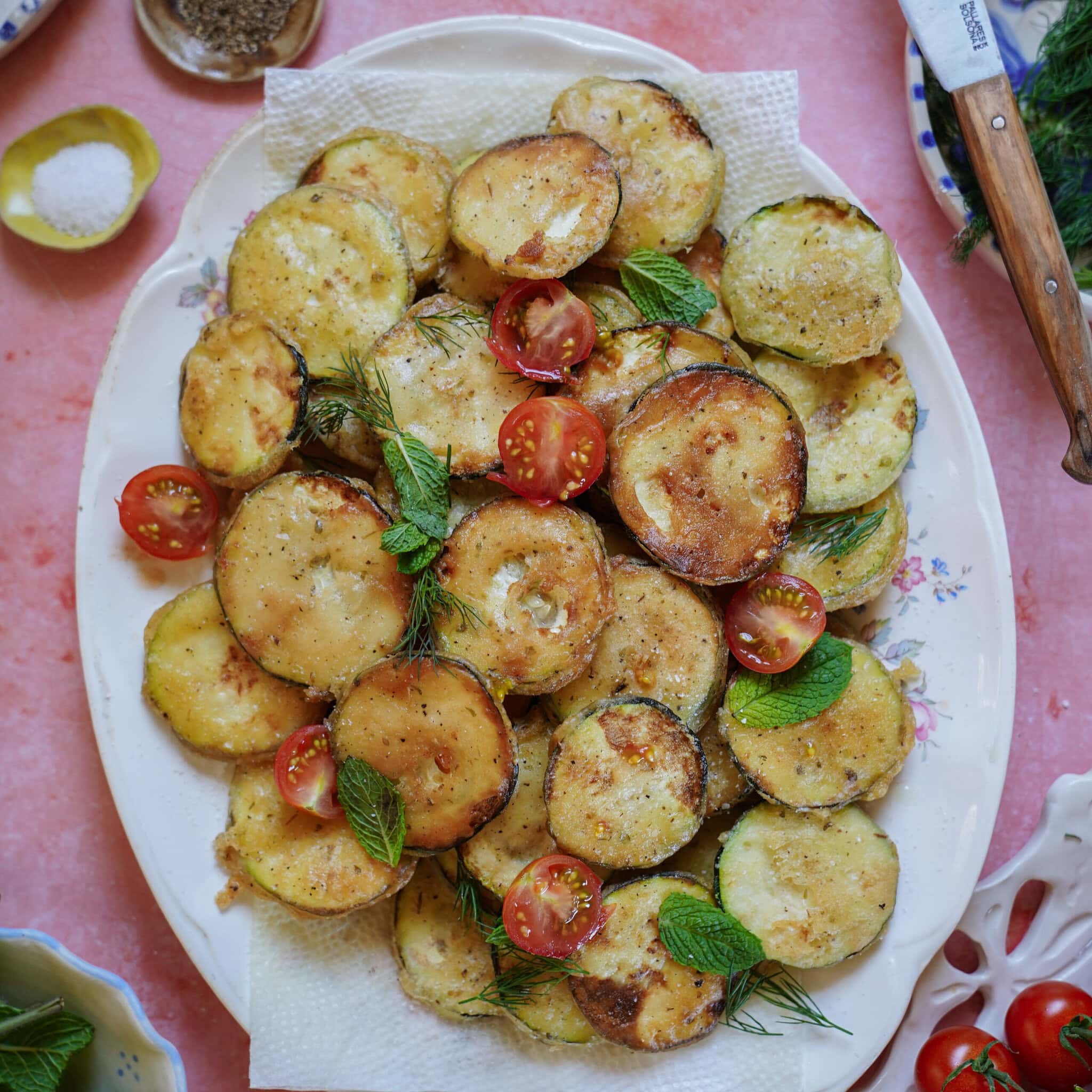 A platter of golden, fried zucchini slices garnished with fresh mint leaves, dill, and halved cherry tomatoes, placed on a pink surface with a small bowl of salt and a knife nearby.