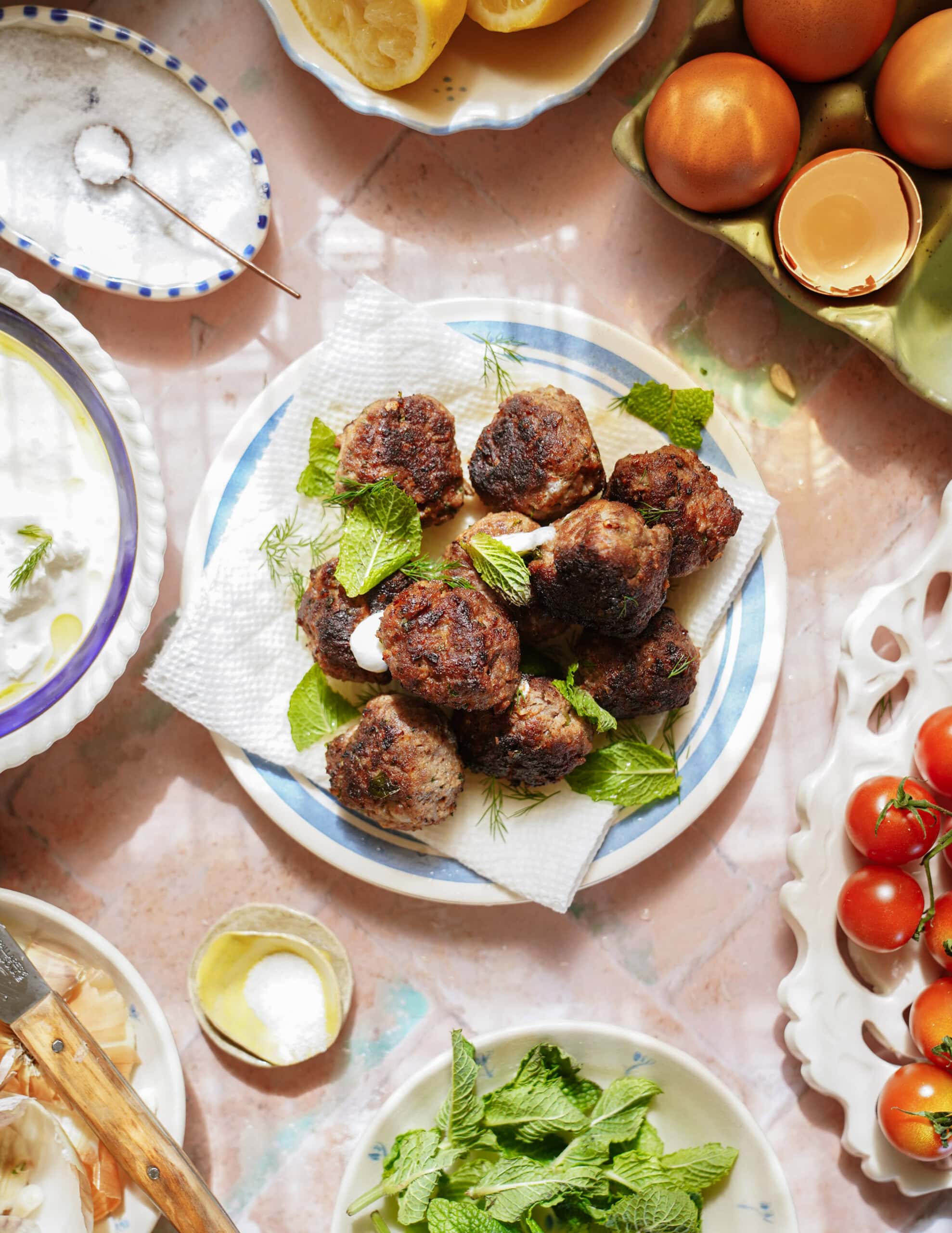 A plate of greek meatballs garnished with fresh mint and herbs sits on a table surrounded by eggs, tomatoes, yogurt sauce, salt, lemon, and mint leaves, creating a vibrant food scene.