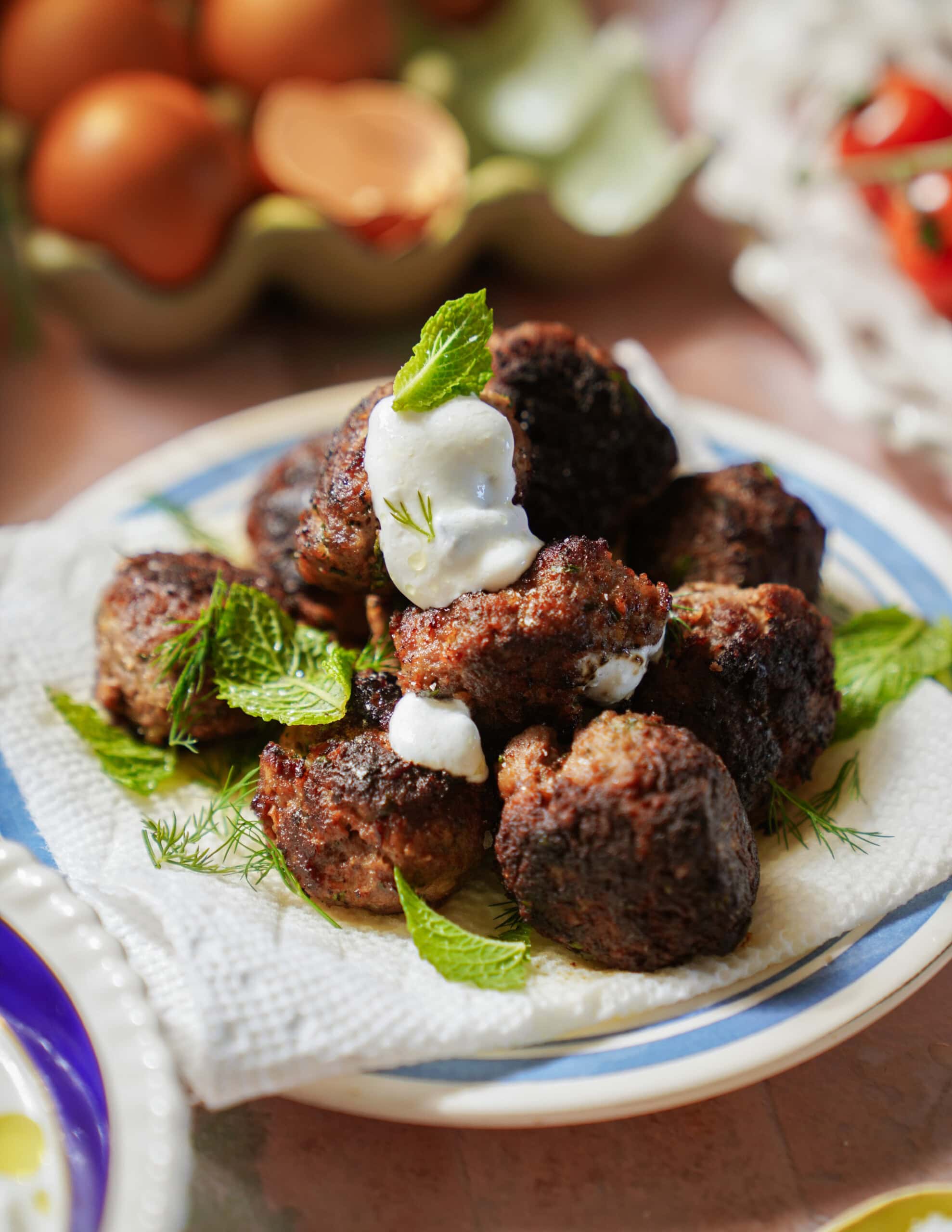 A plate of browned greek meatballs garnished with fresh mint leaves and topped with a dollop of white sauce, placed on a napkin, with eggs and herbs visible in the background.