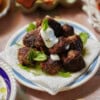 A plate of browned greek meatballs garnished with fresh mint and dill, topped with a dollop of white yogurt sauce, sitting on a paper towel-lined plate.