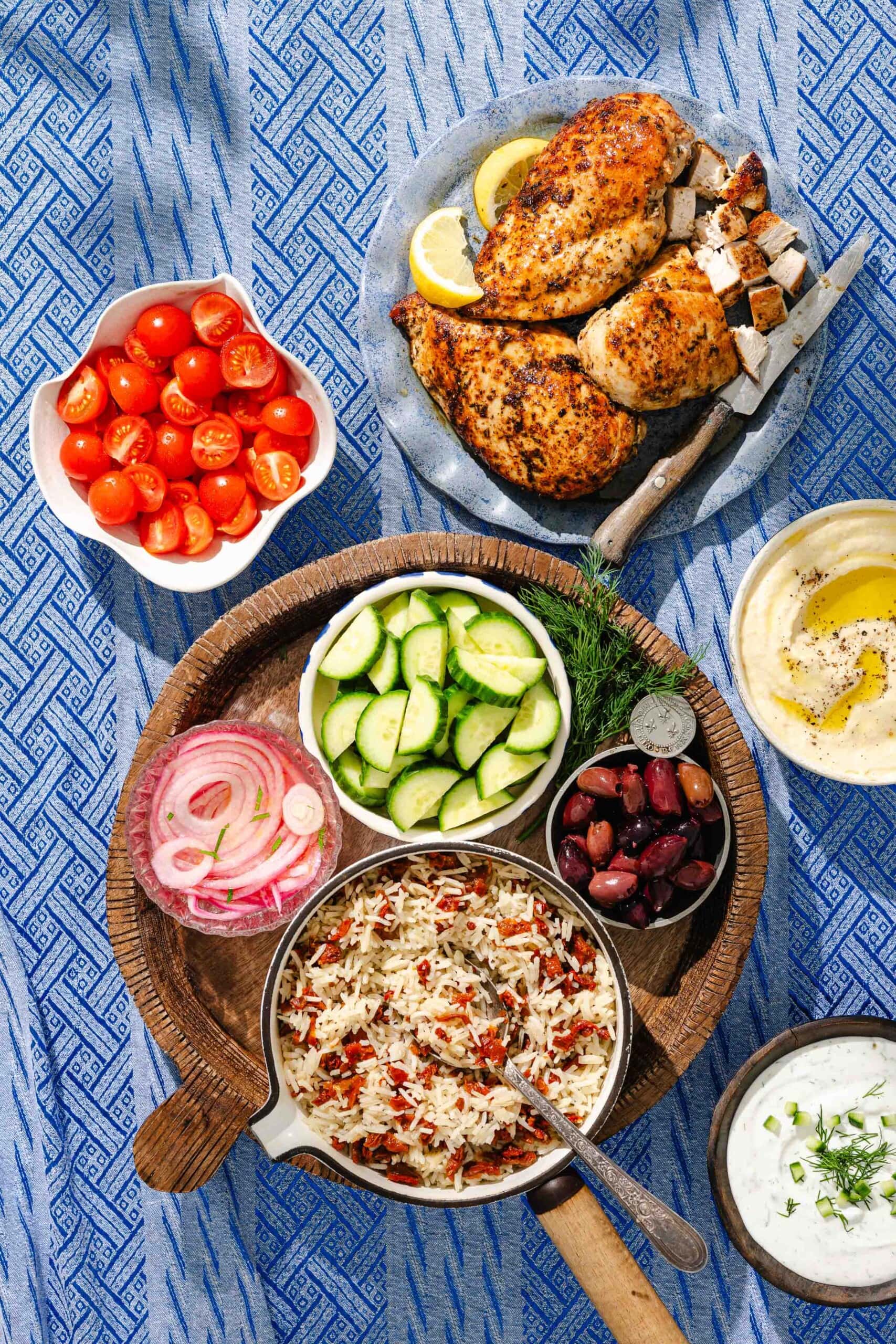 Greek-inspired meal on a blue patterned tablecloth with grilled chicken, sliced cucumbers, cherry tomatoes, hummus, olives, rice pilaf with vegetables, pickled onions, and tzatziki sauce.