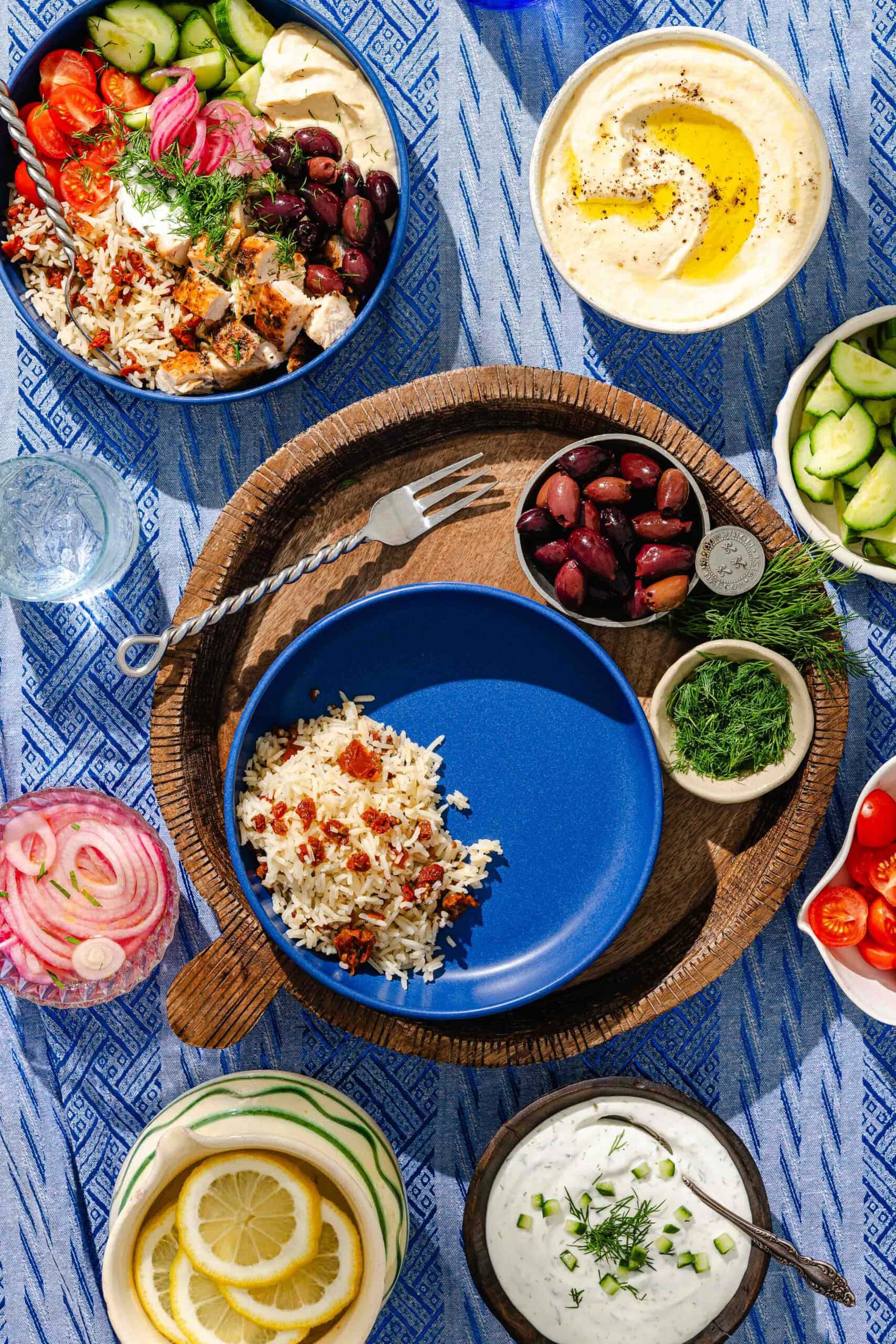 A top-down view of a Mediterranean meal with rice, grilled vegetables, olives, hummus, tzatziki, cucumbers, lemon slices, fresh herbs, tomatoes, and pickled onions arranged on a blue patterned tablecloth.