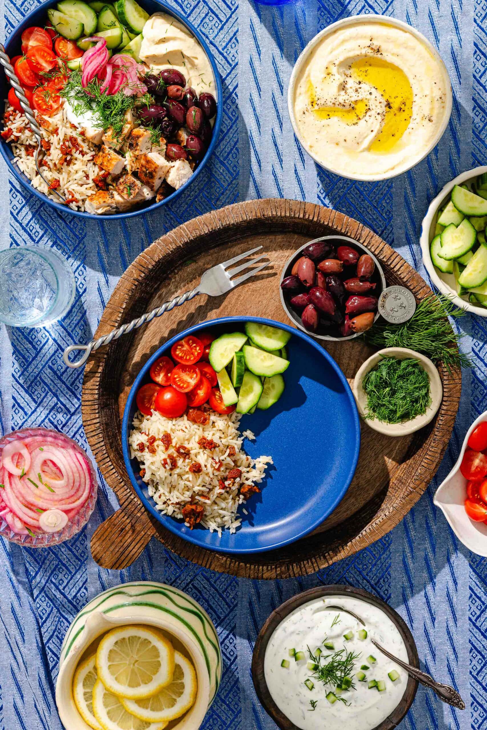 A colorful Mediterranean meal on a blue patterned tablecloth, featuring bowls of rice, cherry tomatoes, cucumbers, olives, tzatziki, hummus drizzled with oil, lemon slices, dill, and pickled onions.