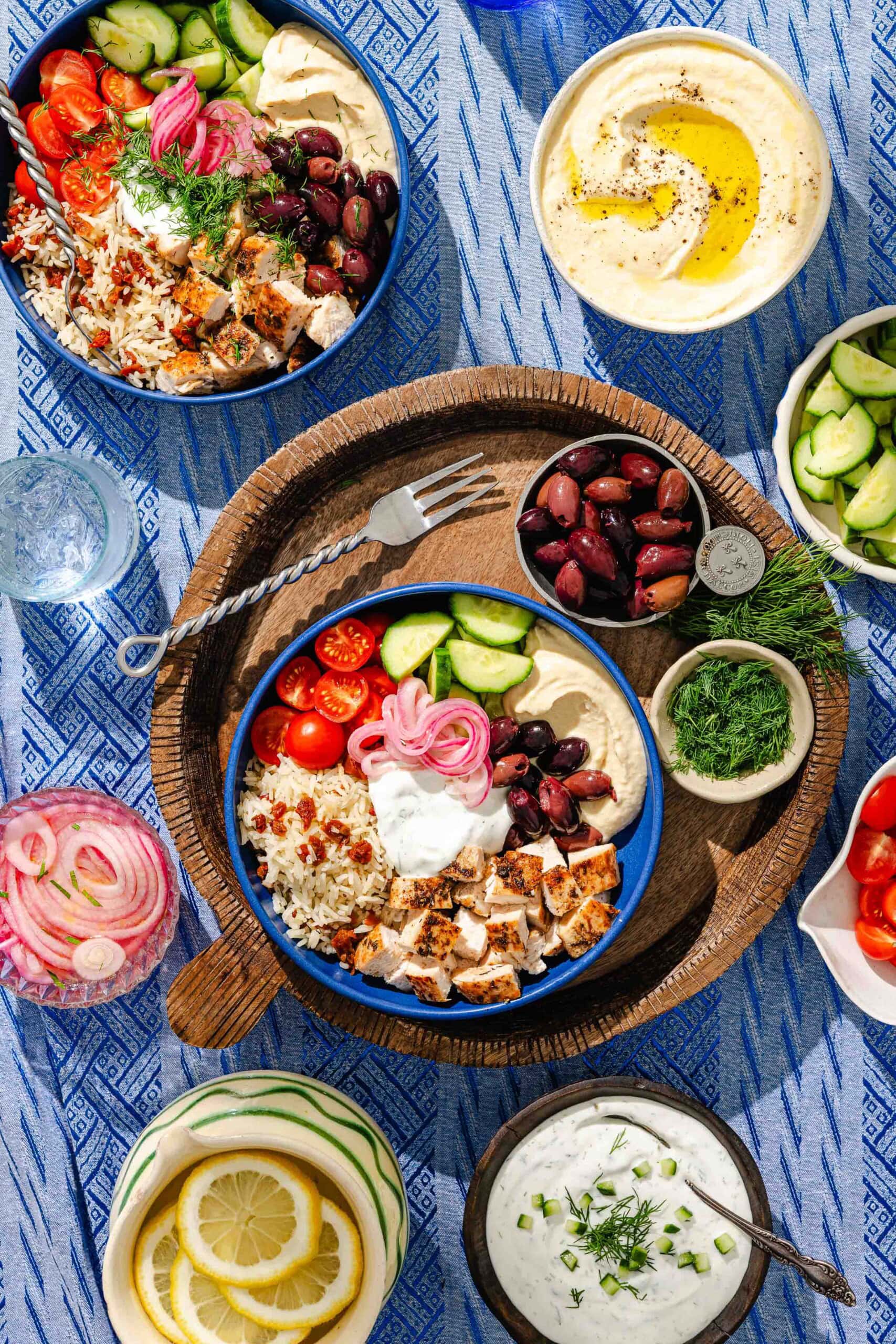 Overhead view of two bowls with rice, grilled chicken, tomatoes, cucumbers, olives, pickled onions, and hummus, surrounded by small bowls of tzatziki, lemon, olives, fresh dill, cucumbers, and tomatoes on a blue patterned tablecloth.